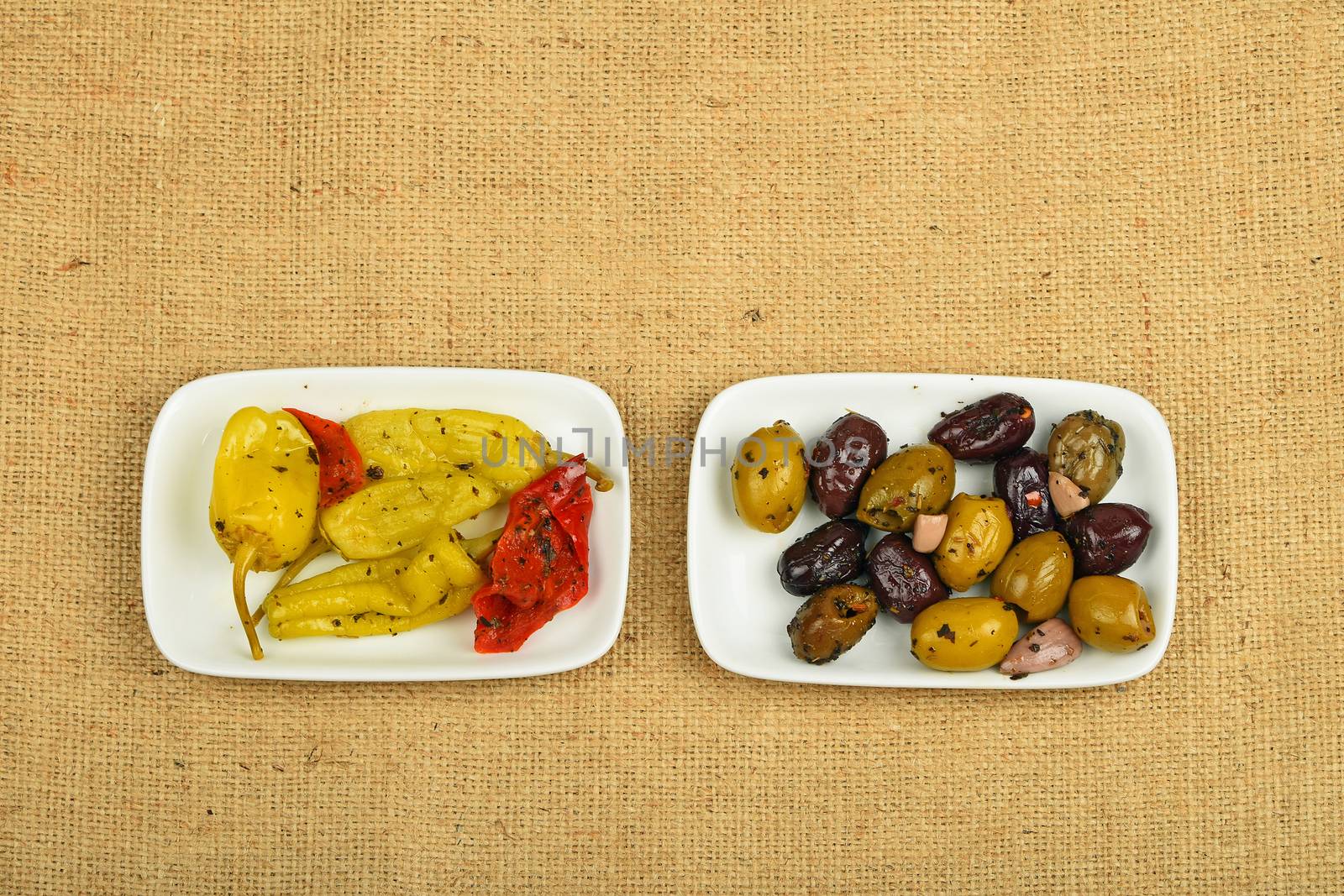 Mediterranean snack of red and green olives and pickled green pepper in two white plates on burlap jute canvas