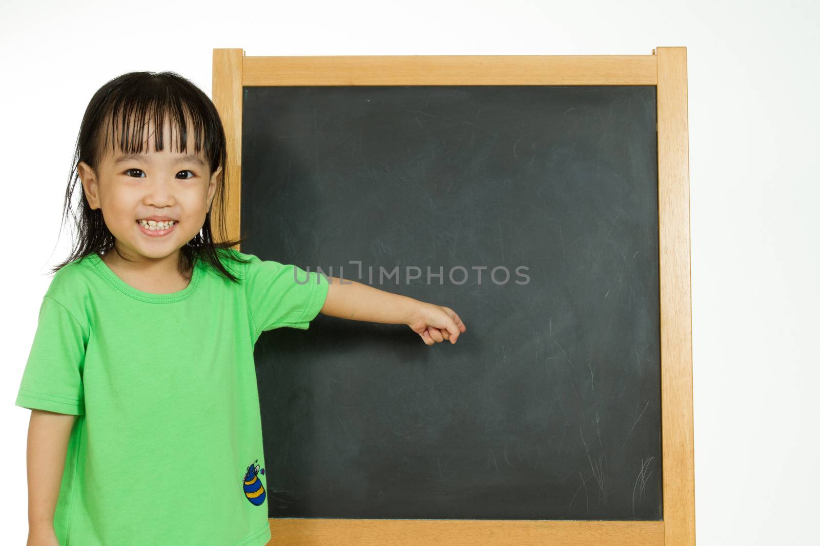 Happy cute Asian Chinese toddler girl standing beside a blank blackboard at home, preschool, daycare or kindergarten in plain white isolated background.