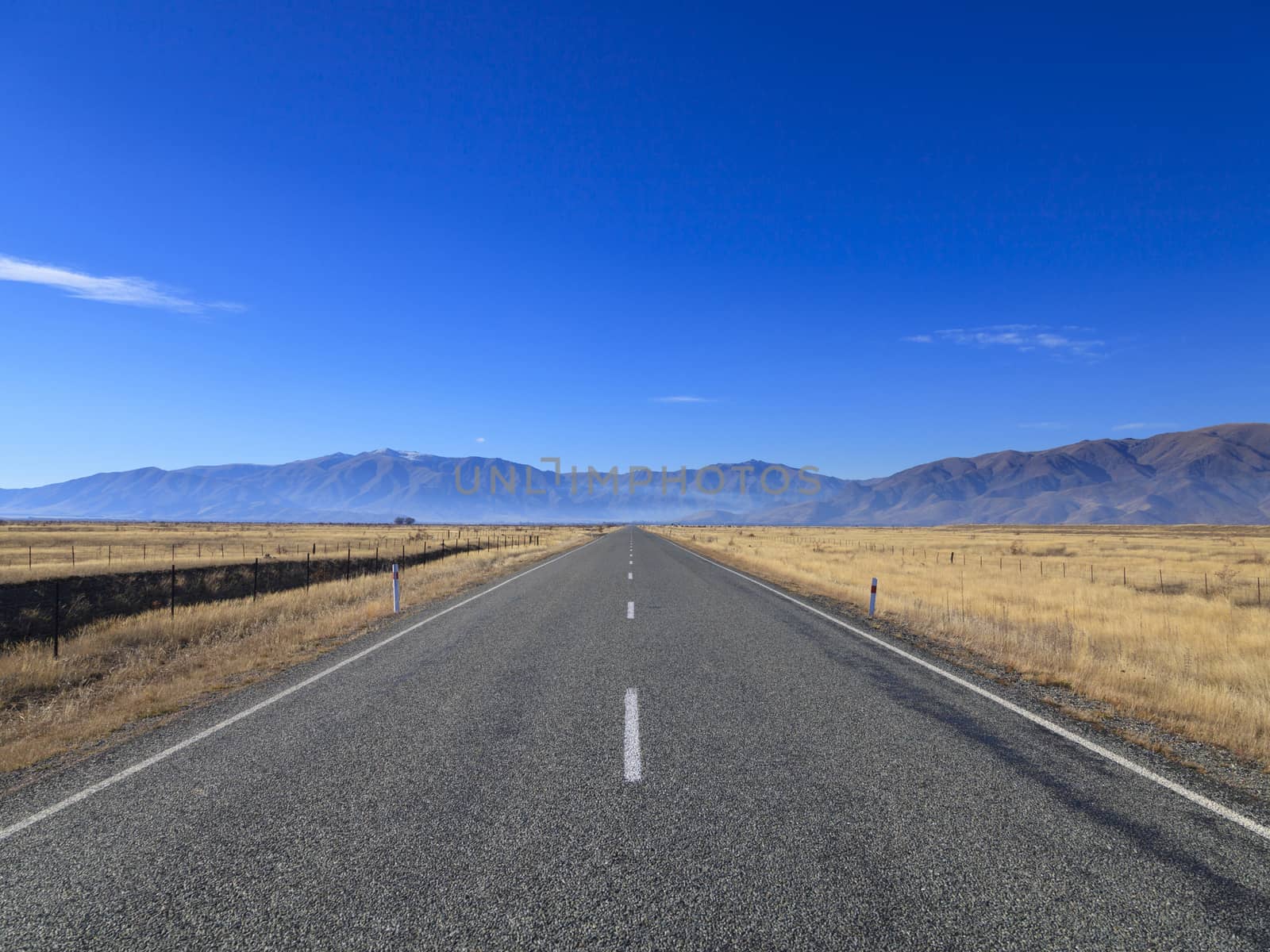 Straight empty Country Road leading to Mountains lining the horizon on sunny day, Otago region, South Island, New Zealand.