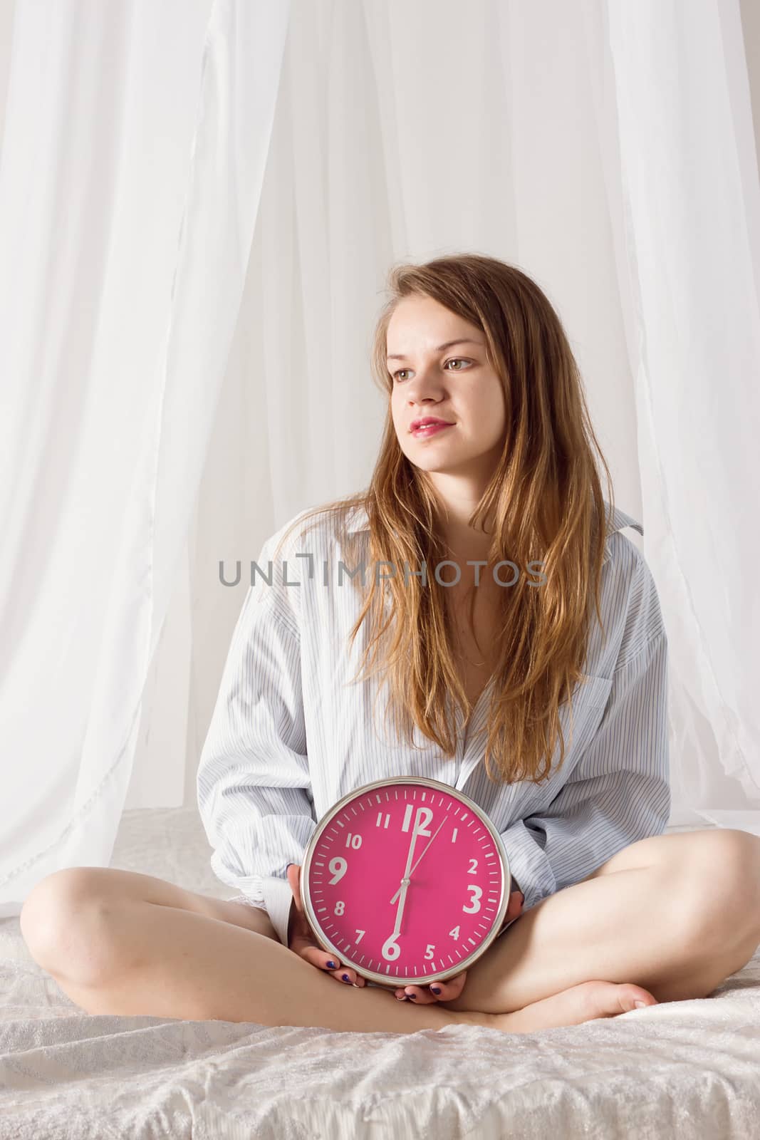 girl is sitting on the bed with big pink clock. 6 a.m