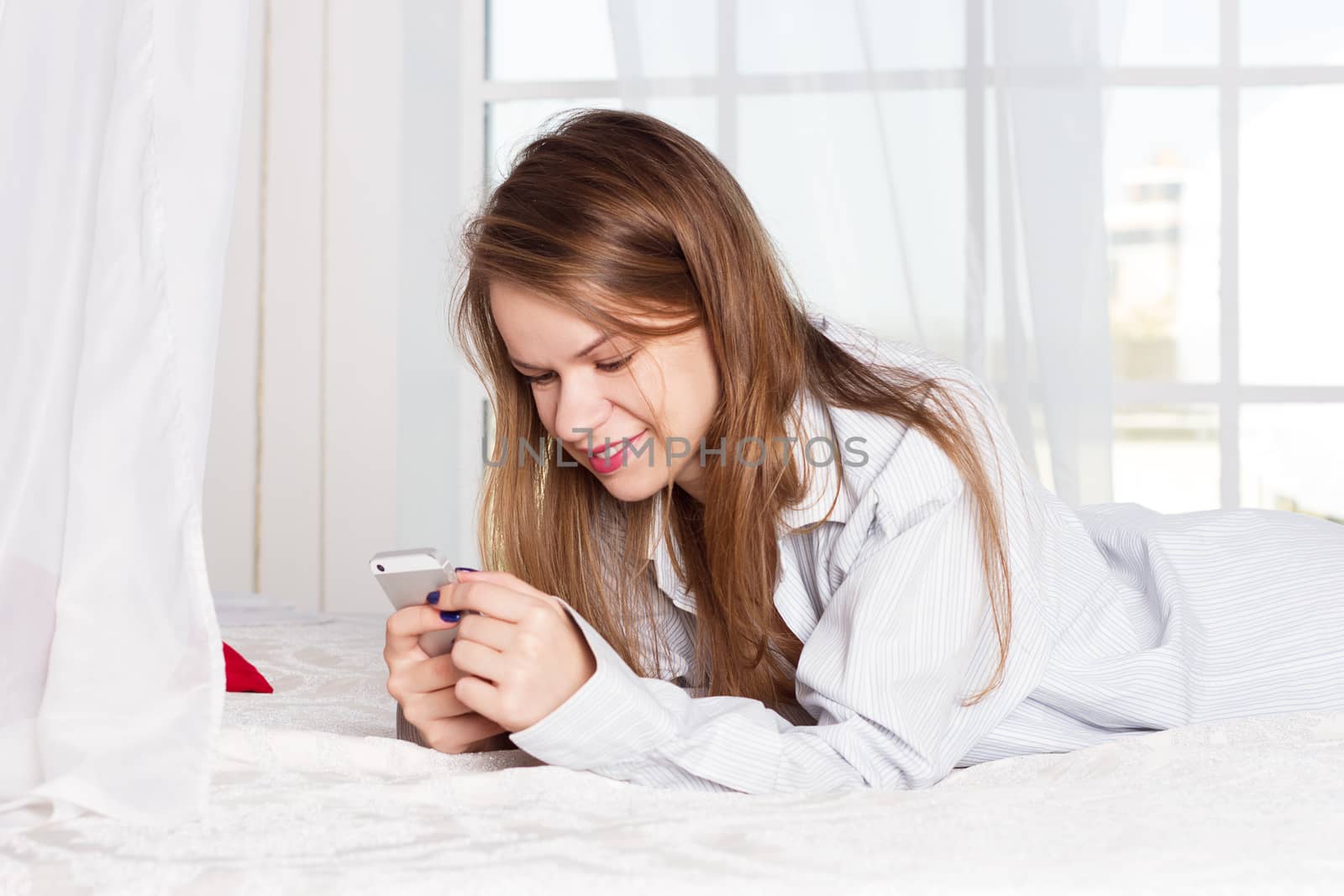 girl lies in a man's shirt on the bed with a smartphone in the hands of