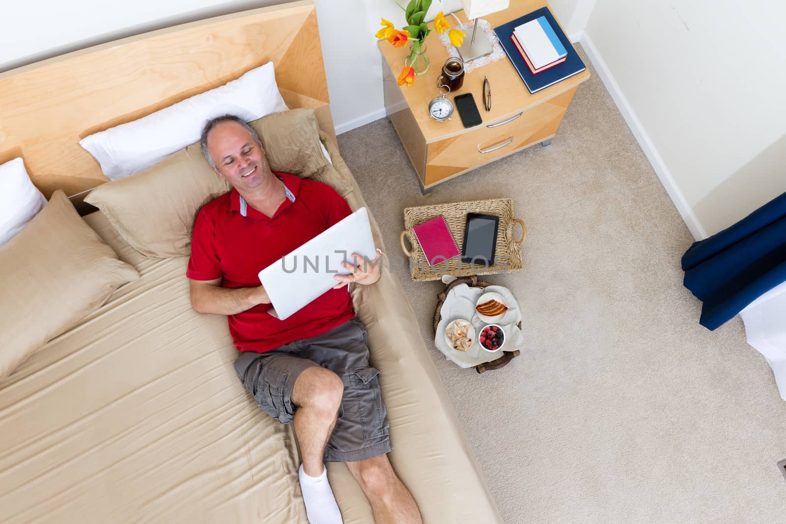 High Angle View of Smiling Mature Man Lying on Bed with Laptop in Hotel Room with Remnants of Breakfast Tray and Personal Effects on Floor
