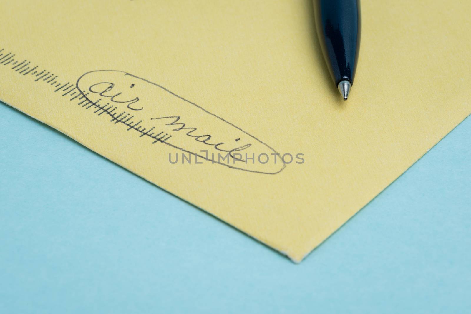 A yellow envenlope with the words "air mail" written by hand in pen with the pen lying on top and a blue background.