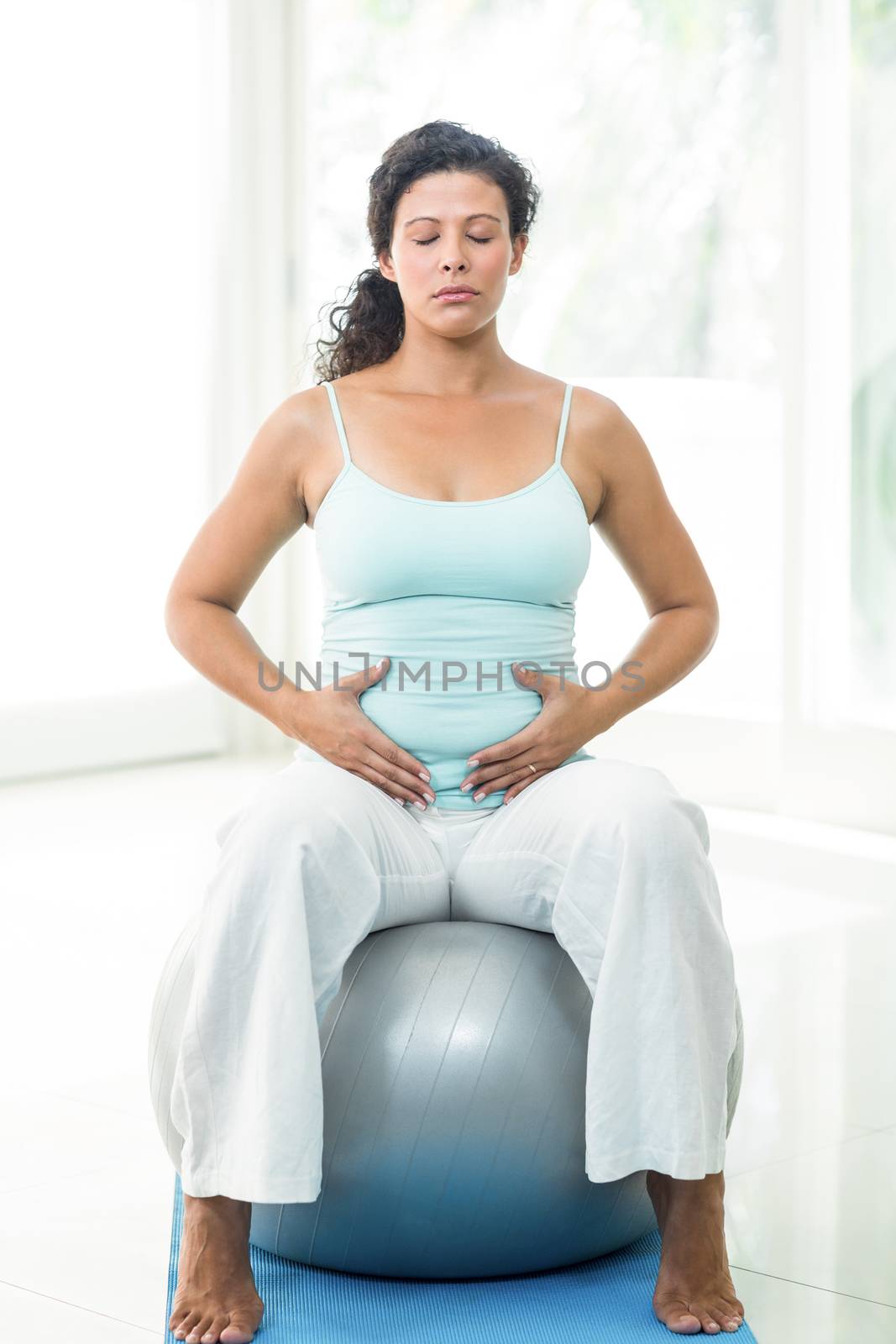 Full length of pregnant woman with eyes closed touching her belly while sitting on exercise ball in fitness studio