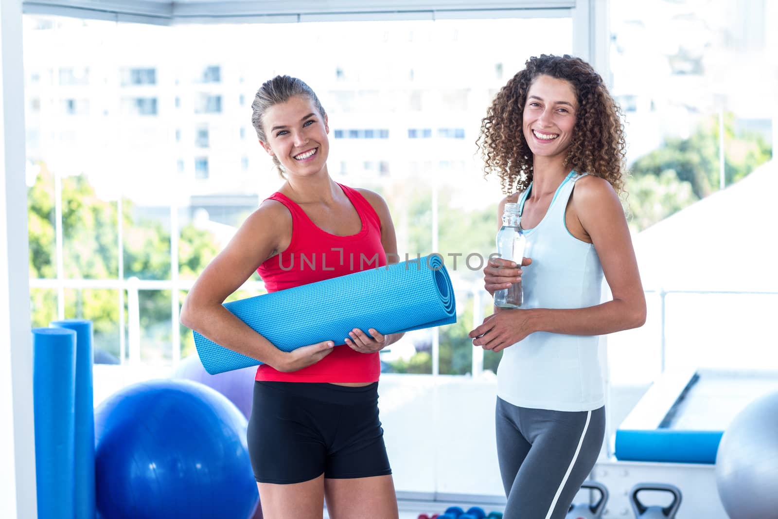 Portrait of fit women holding exercise mat and water bottle in fitness studio
