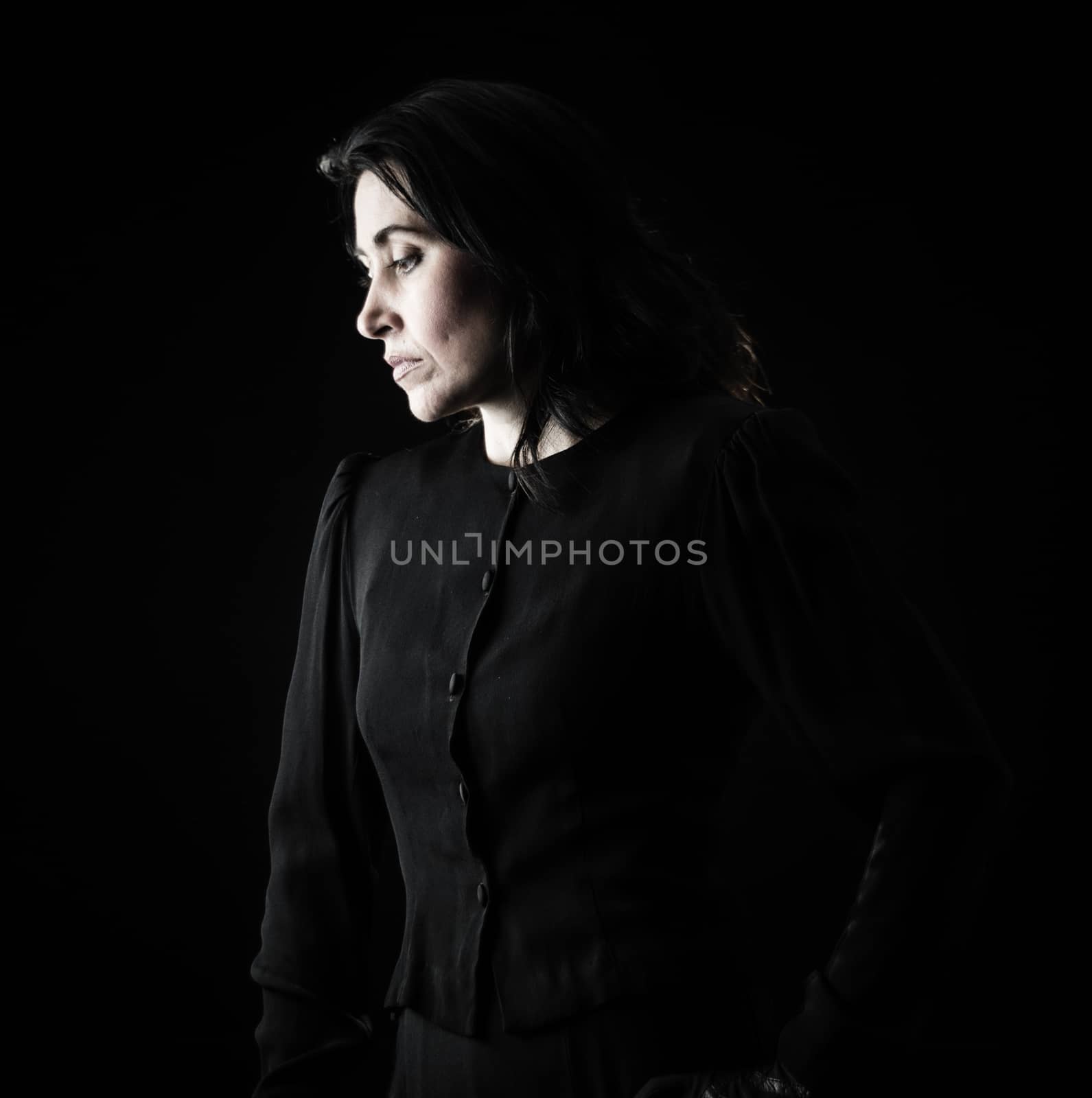 Brunette woman in black standing in front of black backdrop, with a hand on her hip and looking down with a serious, sad expression on her face