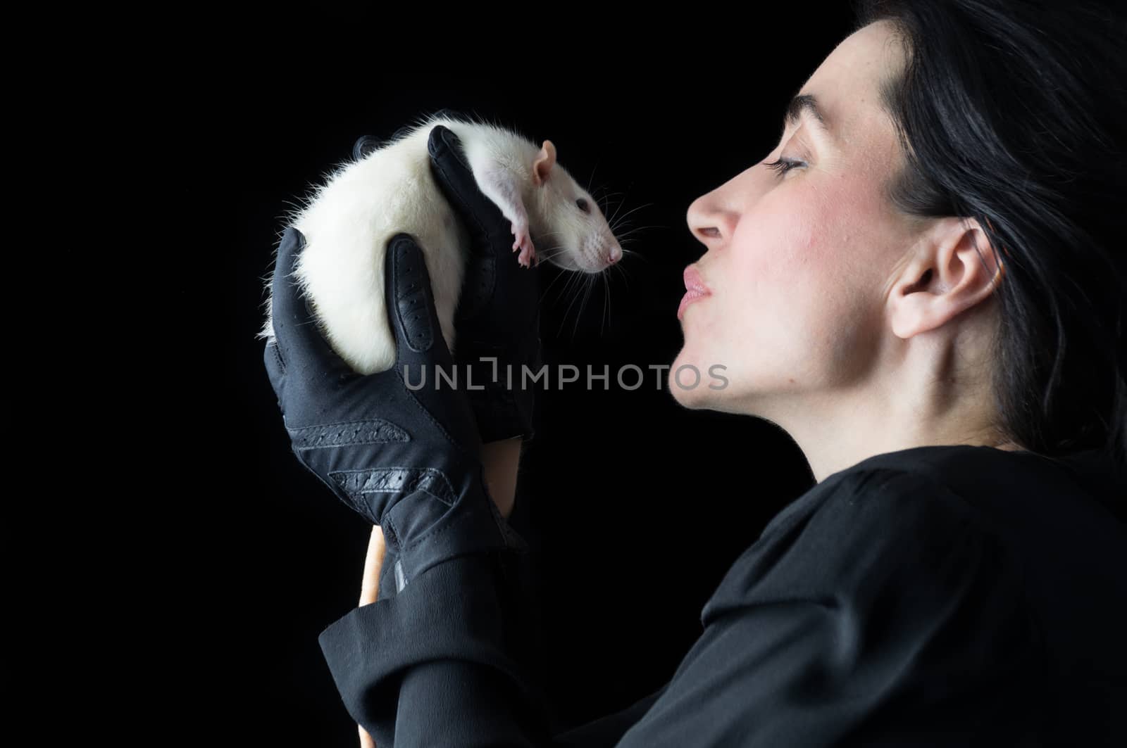 Brunette woman in black standing in front of a black backdrop, holding a white dumbo rat, looking at it with affection and love.