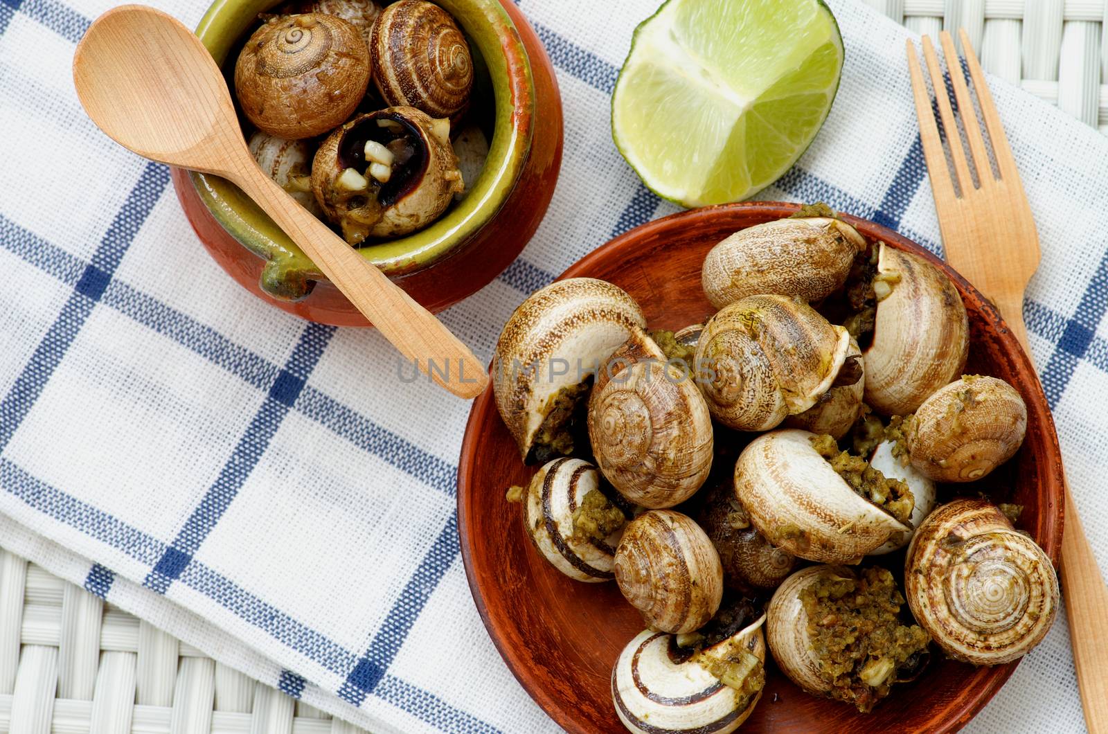 Delicious Escargot with Garlic Butter in Two Plates with Wooden Fork, Wooden Spoon and Lime closeup on Checkered Napkin