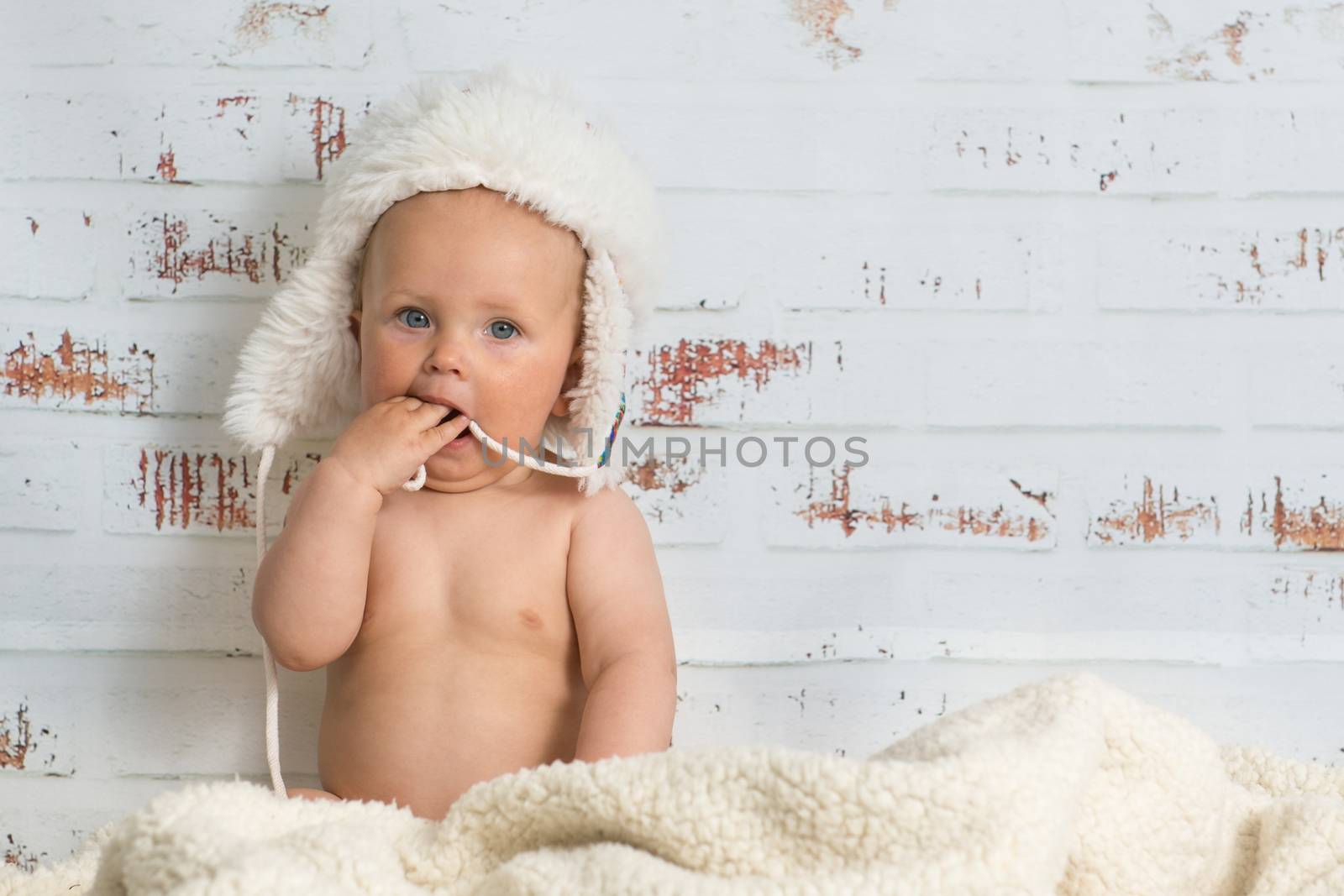 Naked baby girl sitting in a warm interior wearing white cap. Rustic wall in the back.