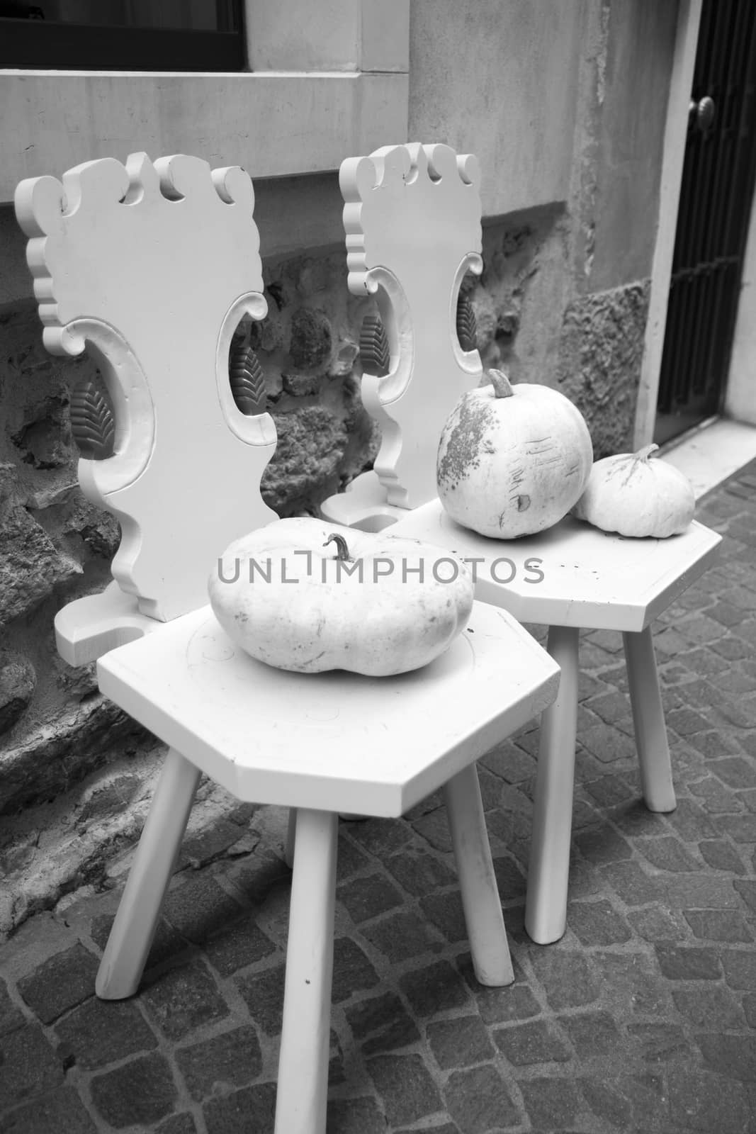 two wooden chairs painted white and two white pumpkins against the wall along a street