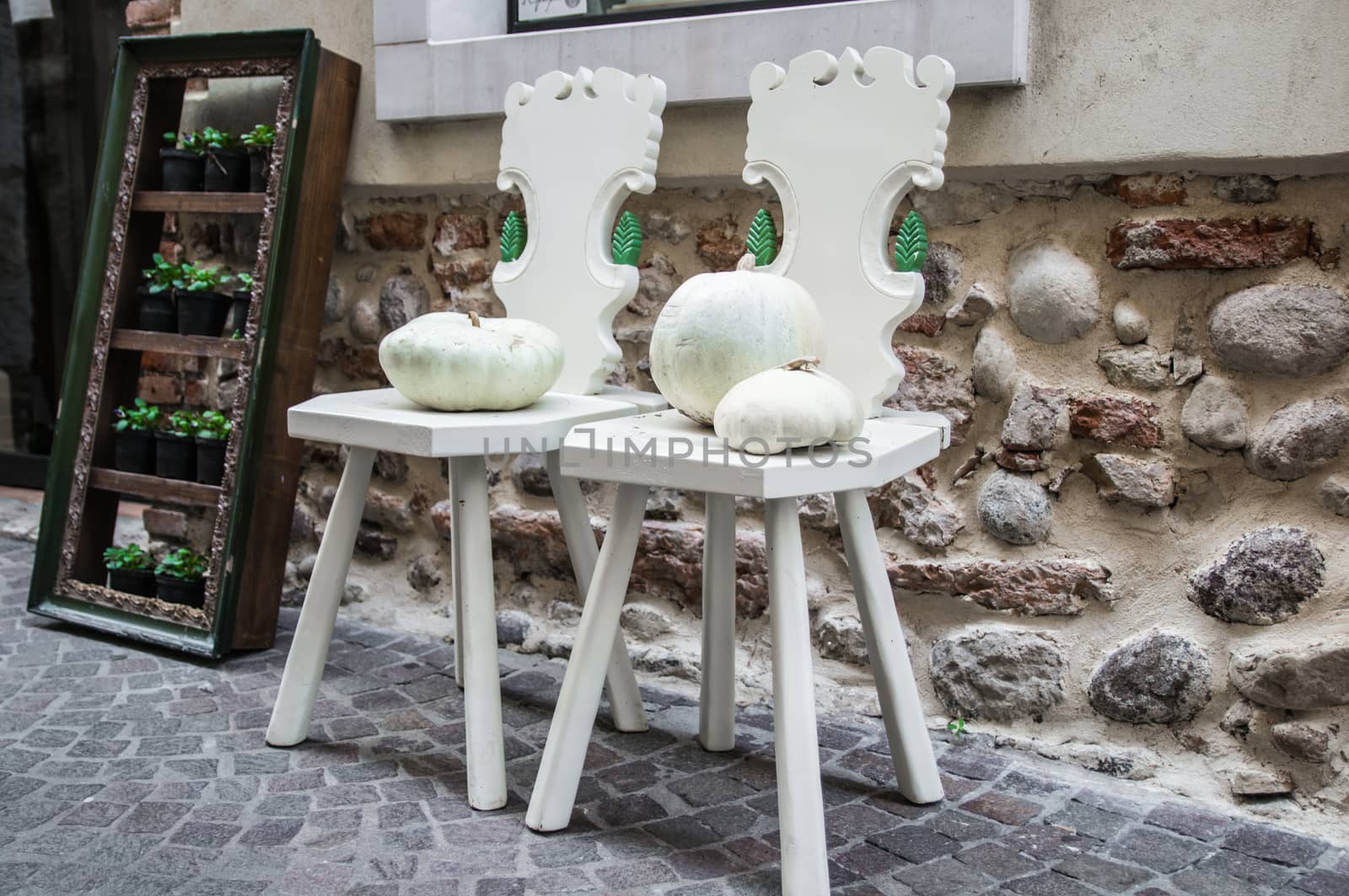 two wooden chairs painted white and two white pumpkins against the wall along a street