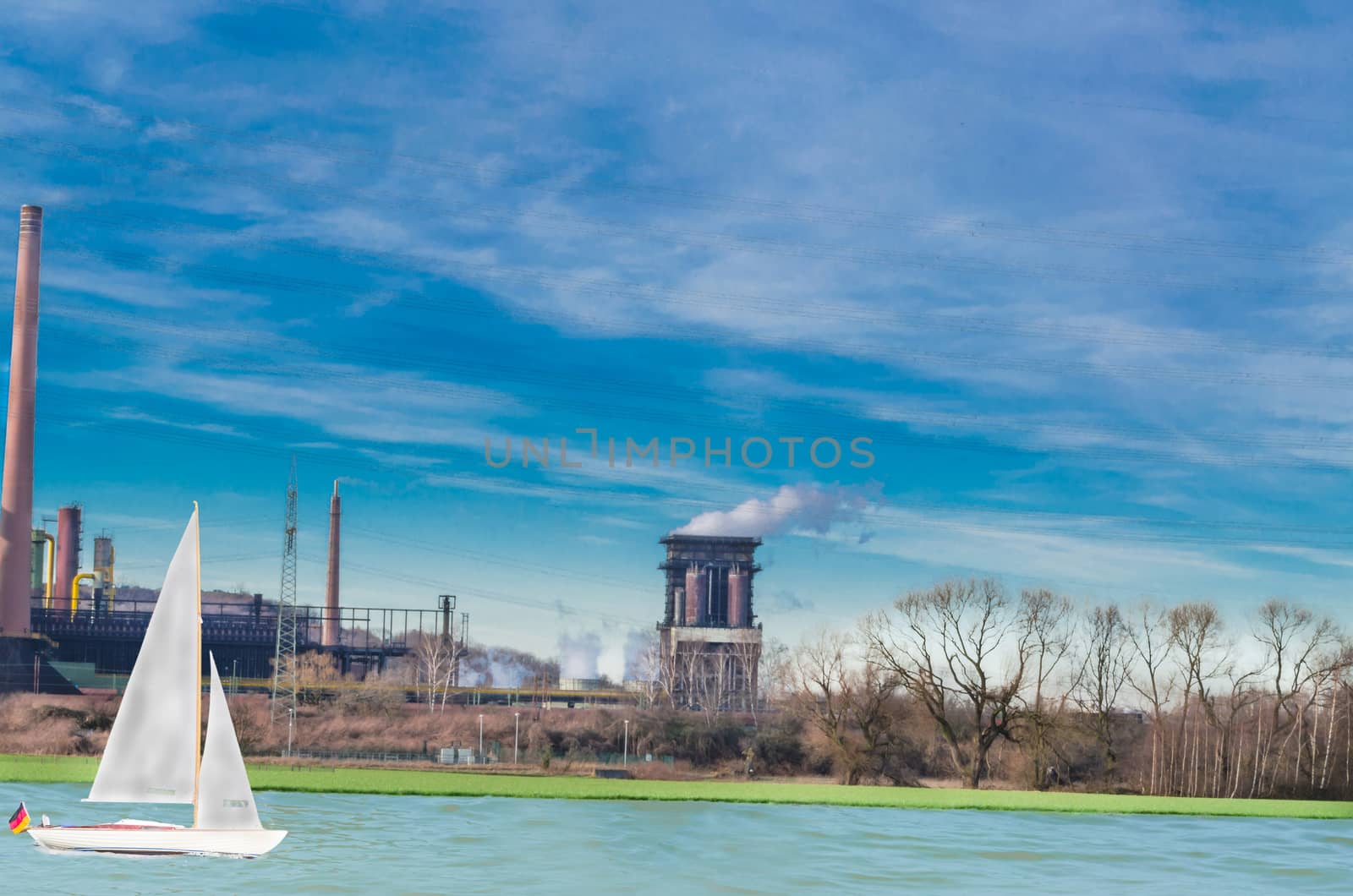 Old power plant on a river against a blue sky. On the water a sail boat. Industry, nature and leisure in harmony.