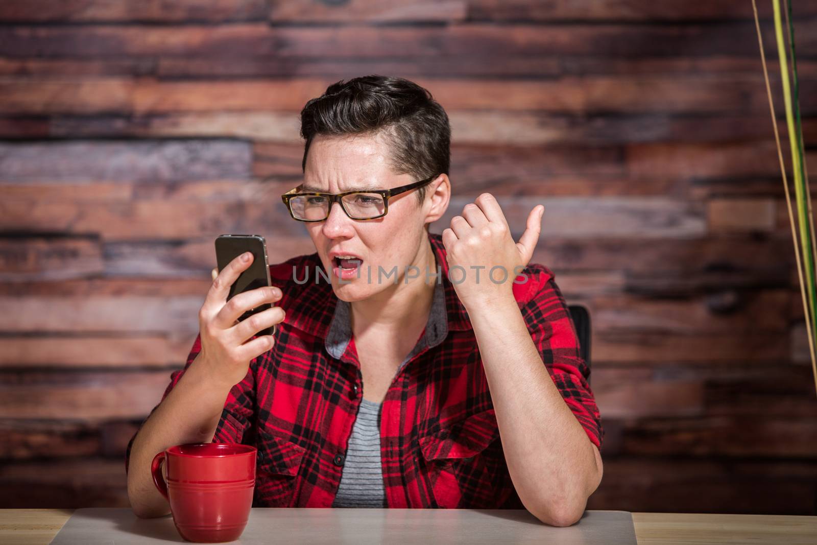 Serious woman wearing flannel shirt looking at smart phone and seated in front of modern desk at hipster office