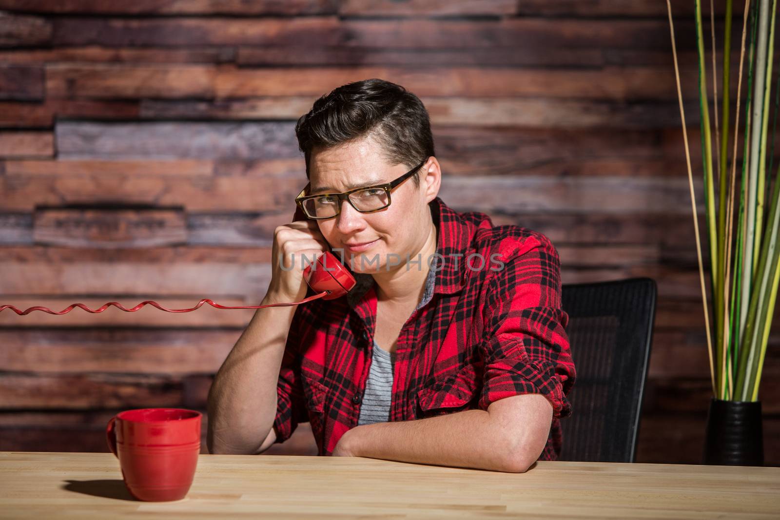 Woman wearing flannel shirt on push-button phone seated in front of modern desk at hipster office