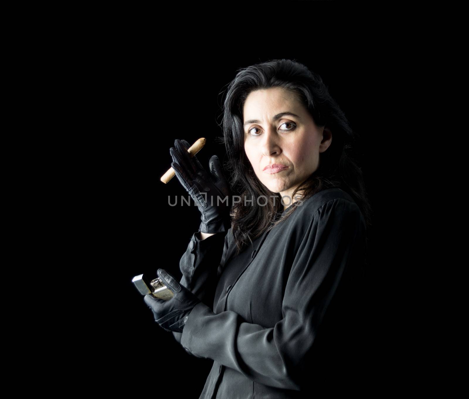 Brunette woman in black dress and black gloves standing in front of a black backdrop, holding a cigar in one hand and a lighter in the other and looking at the camera.