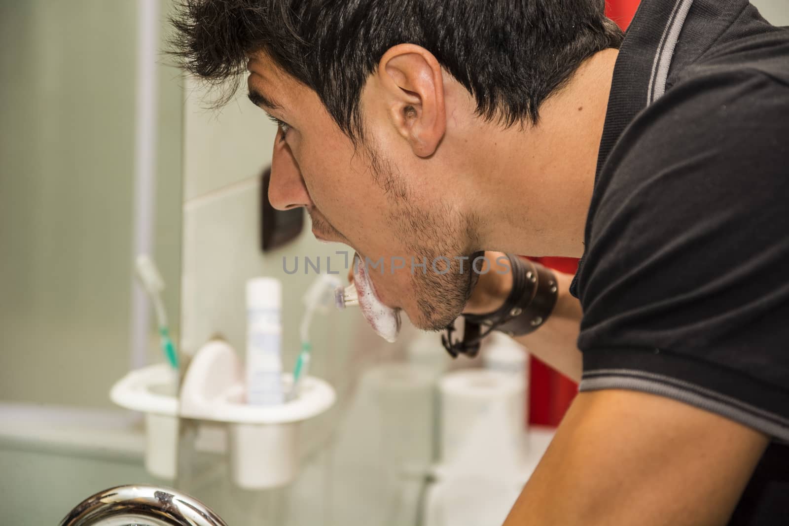 Headshot of attractive young man brushing teeth and cleaning tongue with toothbrush, looking at himself in mirror