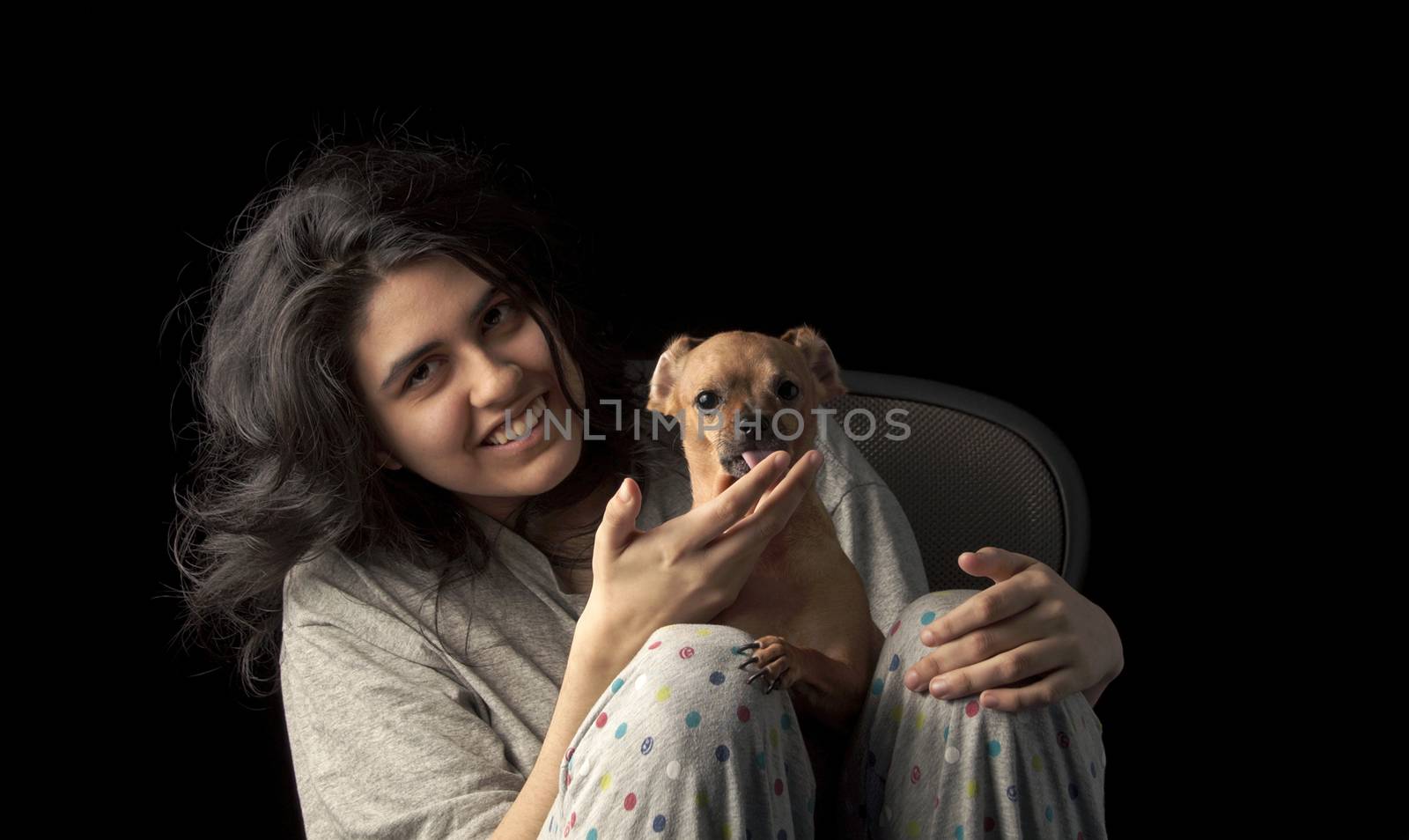 Low key shot of latina teenage girl holding her chihuahua dog and looking at the camera with a smile on her face; the dog's ears are curled back to create a funny appearance and the dog is licking her hand.