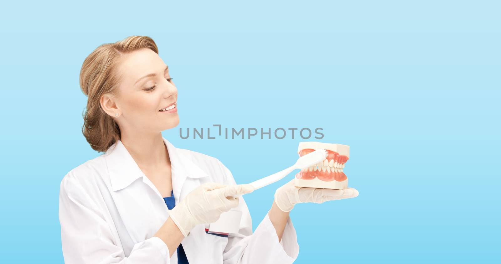 medicine, stomatology, people and hygiene concept - smiling female doctor with toothbrush and jaws model teaching how to brush teeth over blue background