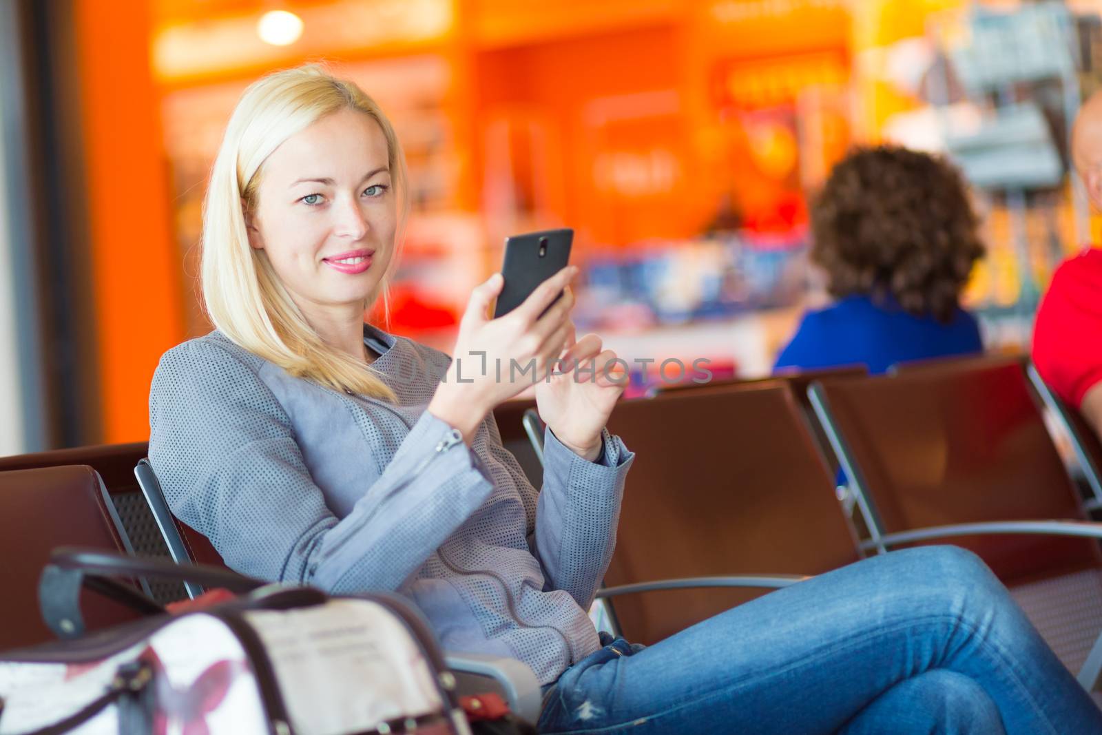 Casual blond young woman using her cell phone while waiting to board a plane at the departure gates. Wireless network hotspot enabling people to access internet conection. Public transport.