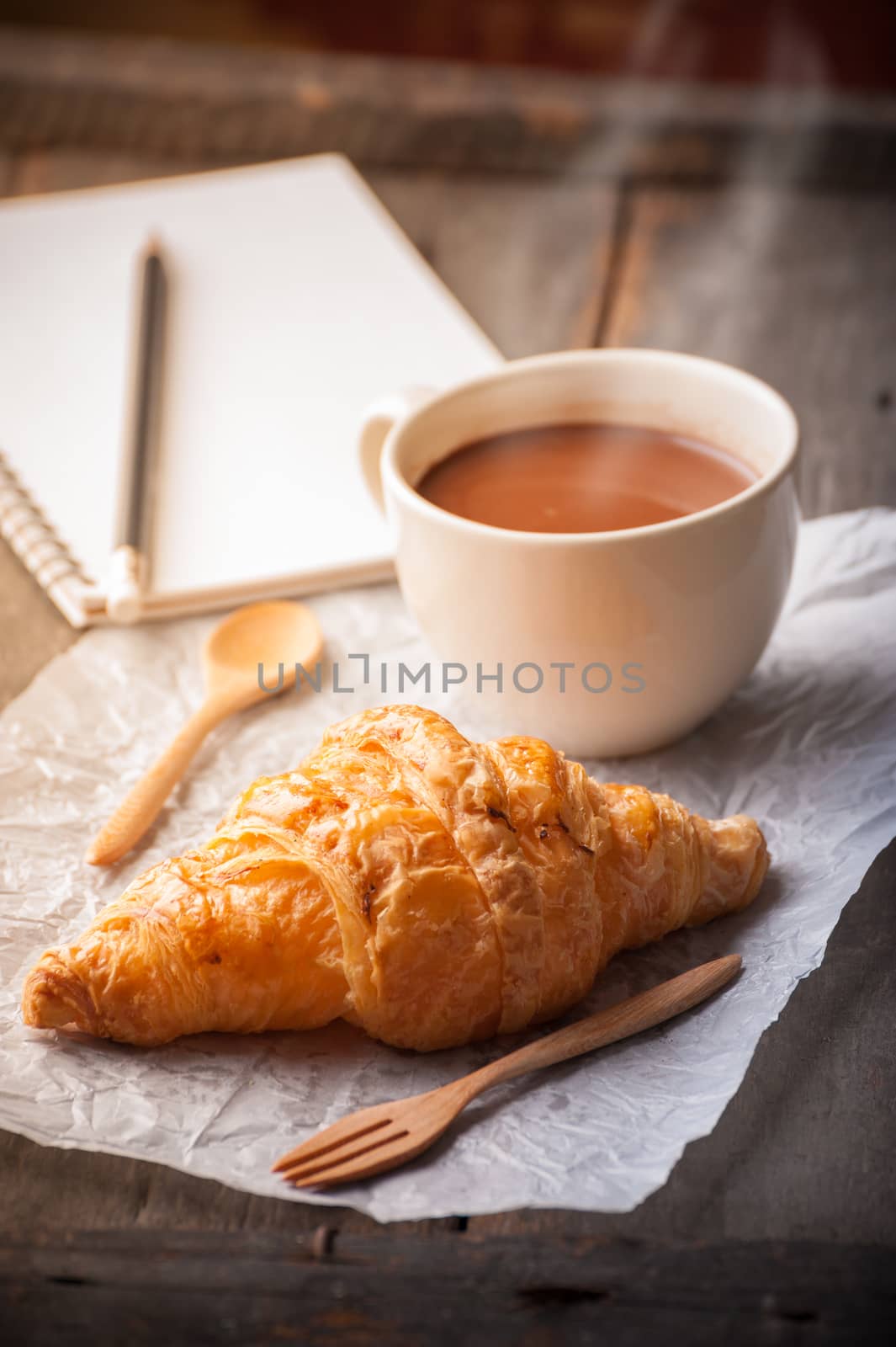 Butter croissant with a cup of hot chocolate and small notebook on wood table with morning scene