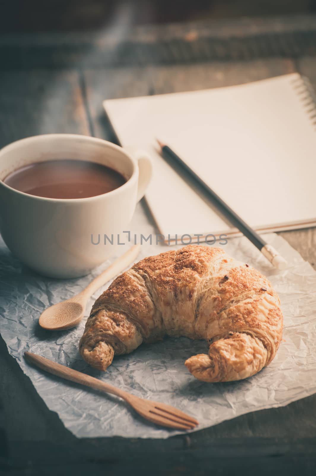 Butter croissant with a cup of hot chocolate and small notebook on wood table in morning time with film filter effect