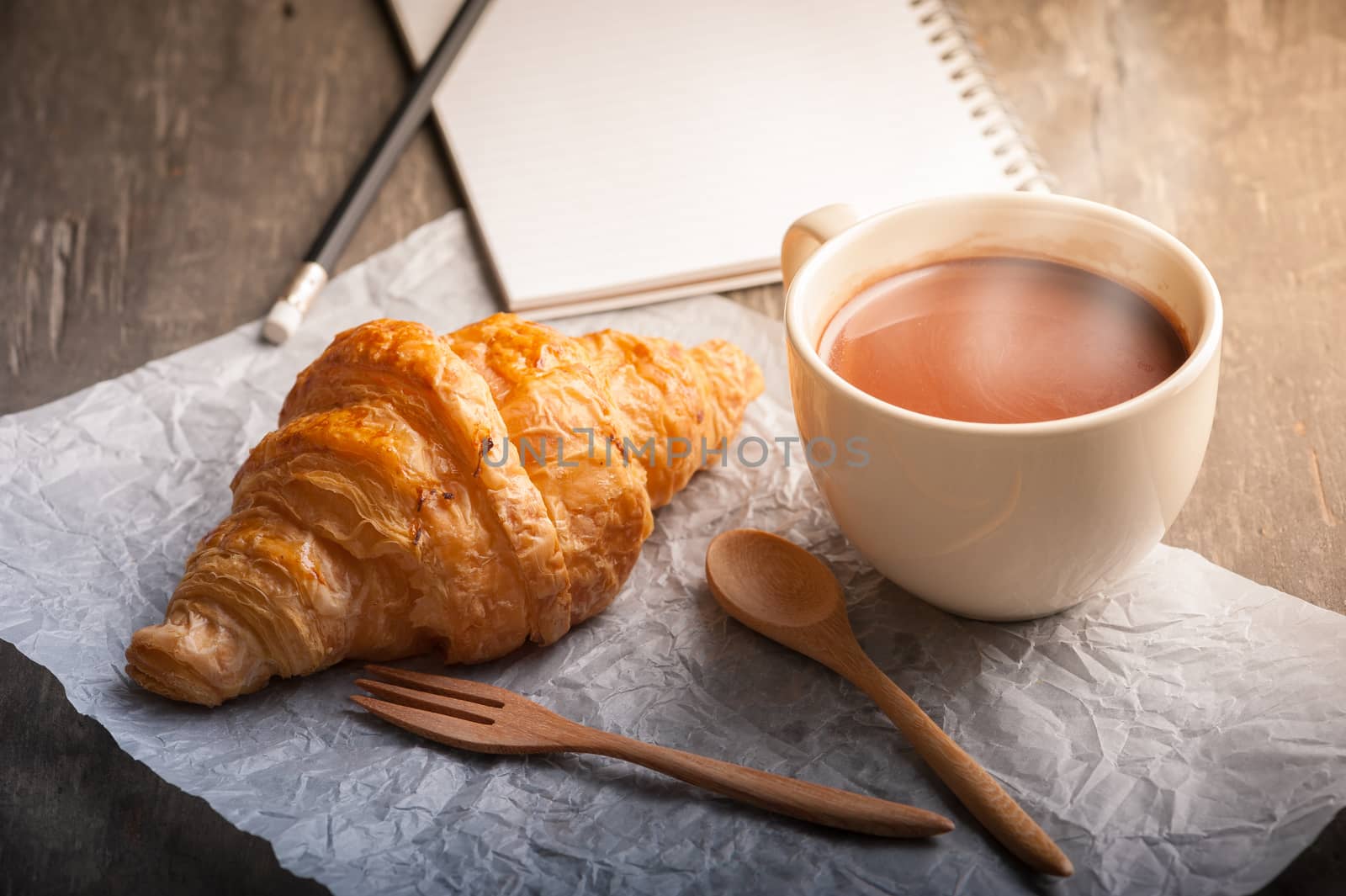 Butter croissant with a cup of hot chocolate and small notebook on wood table with morning scene