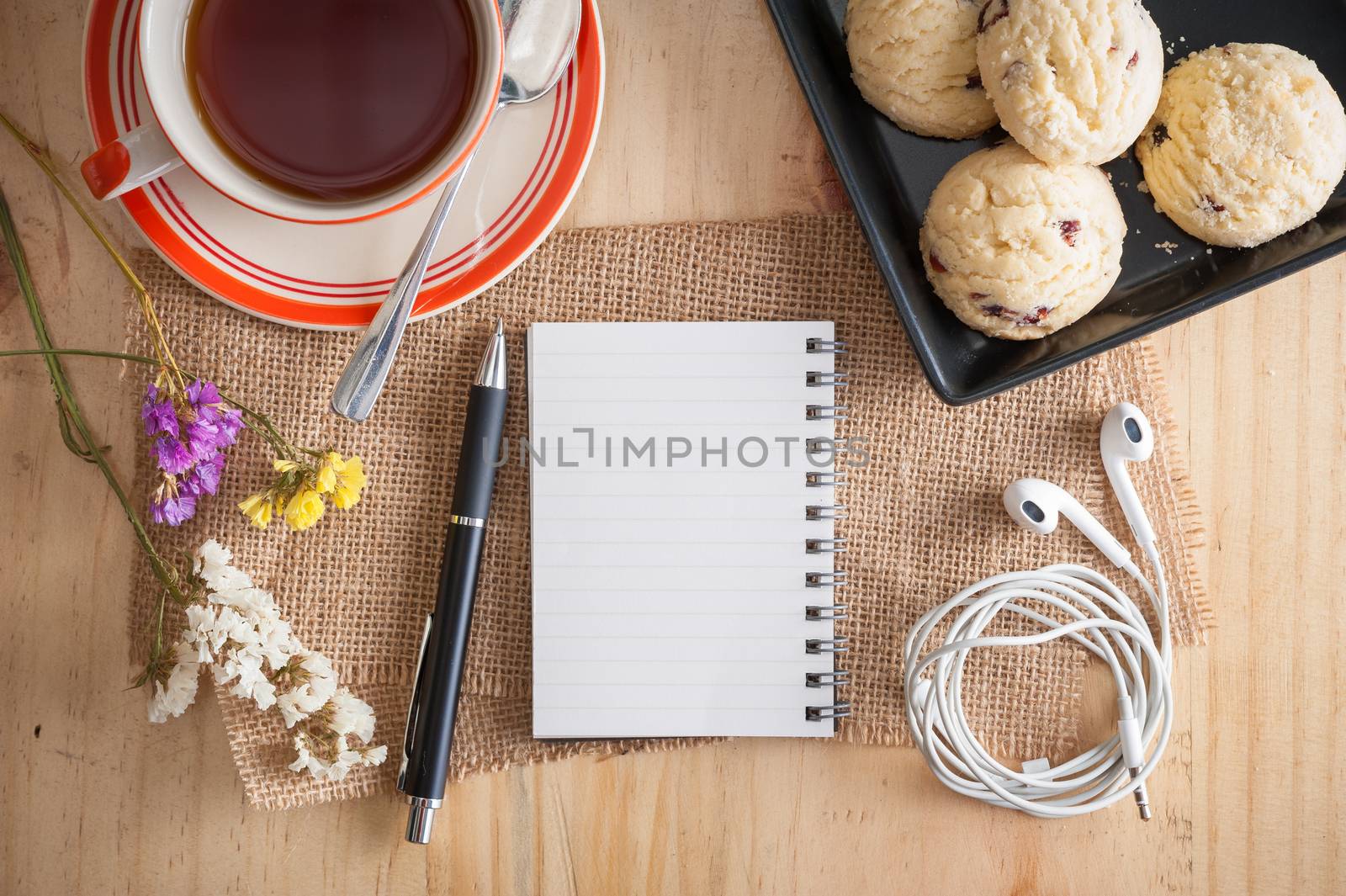 Opened notepad with pen, cookies in black ceramic dish, small earphone, and a cup of hot tea on wood table in cafe with morning scene