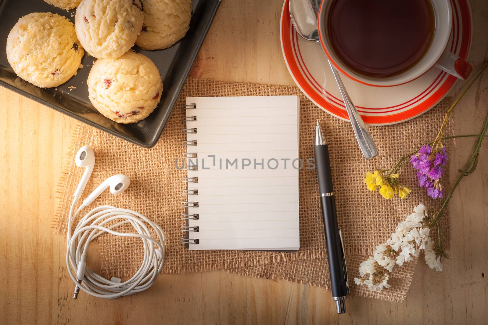 Opened notepad with pen, cookies in black ceramic dish, small earphone, and a cup of hot tea on wood table in cafe with morning light effect