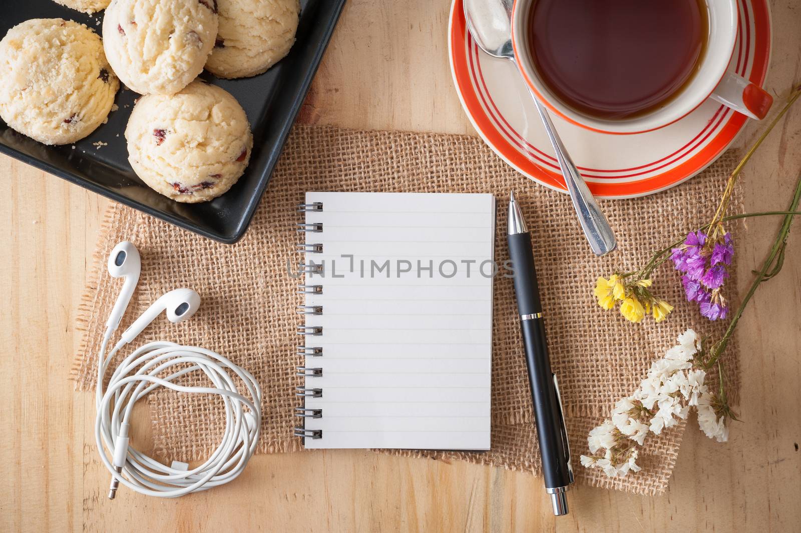 Opened notepad with pen, cookies in black ceramic dish, small earphone, and a cup of hot tea on wood table in cafe with afternoon scene