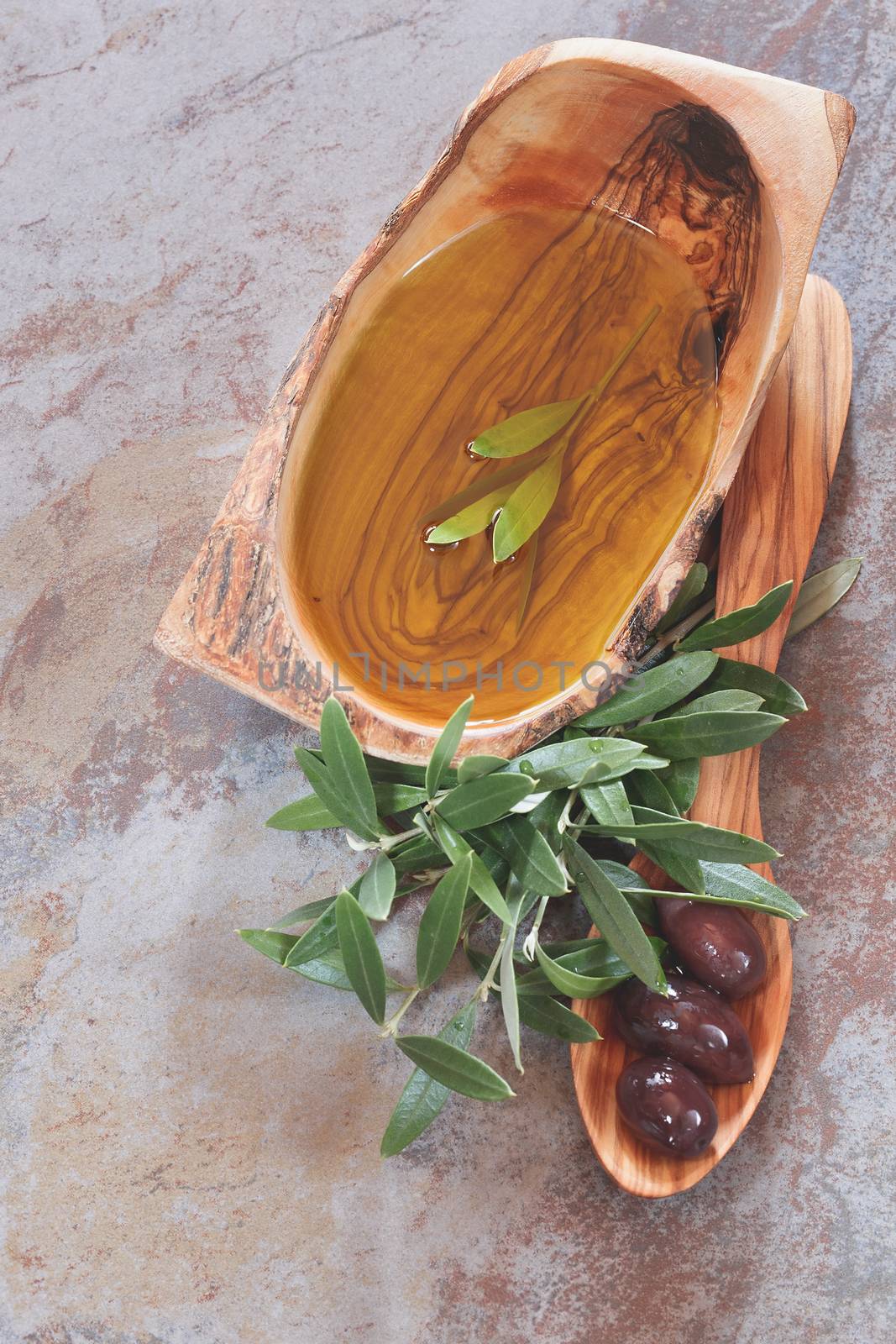 Bowl of olive oil, olive leaves on and around a wooden spoon with black olives . Macro, selective focus, top view