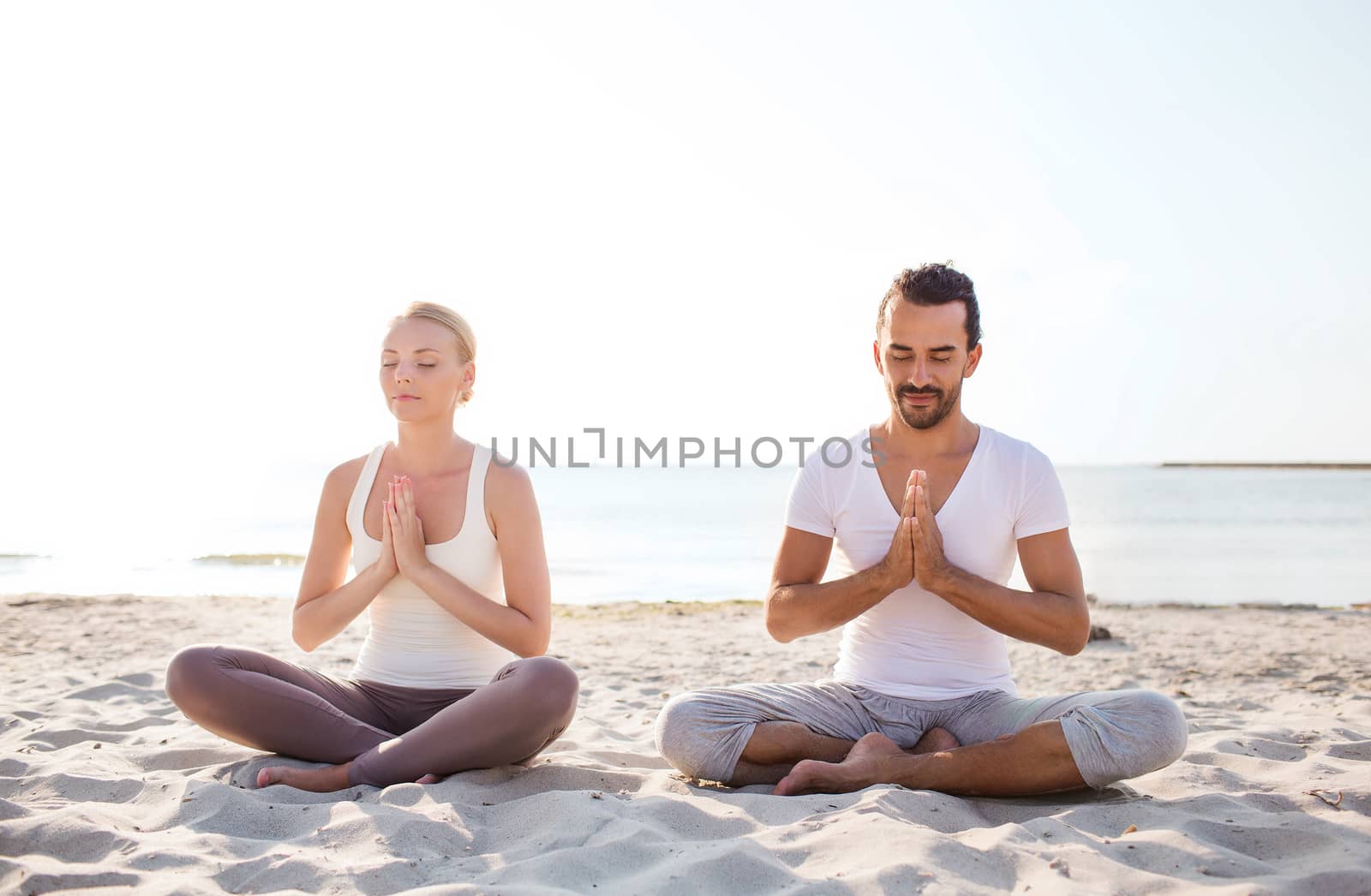 fitness, sport, friendship and lifestyle concept - smiling couple making yoga exercises sitting on sand outdoors