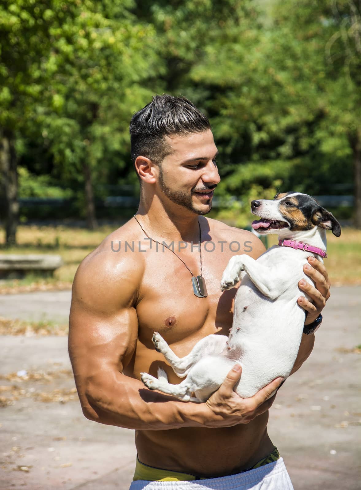 Half Body Shot of a Shirtless Athletic Young Man Carrying One Dog in his Arms and Looking at the Camera.