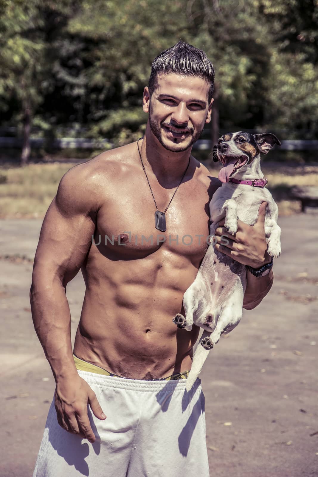 Half Body Shot of a Shirtless Athletic Young Man Carrying One Dog in his Arms and Looking at the Camera.