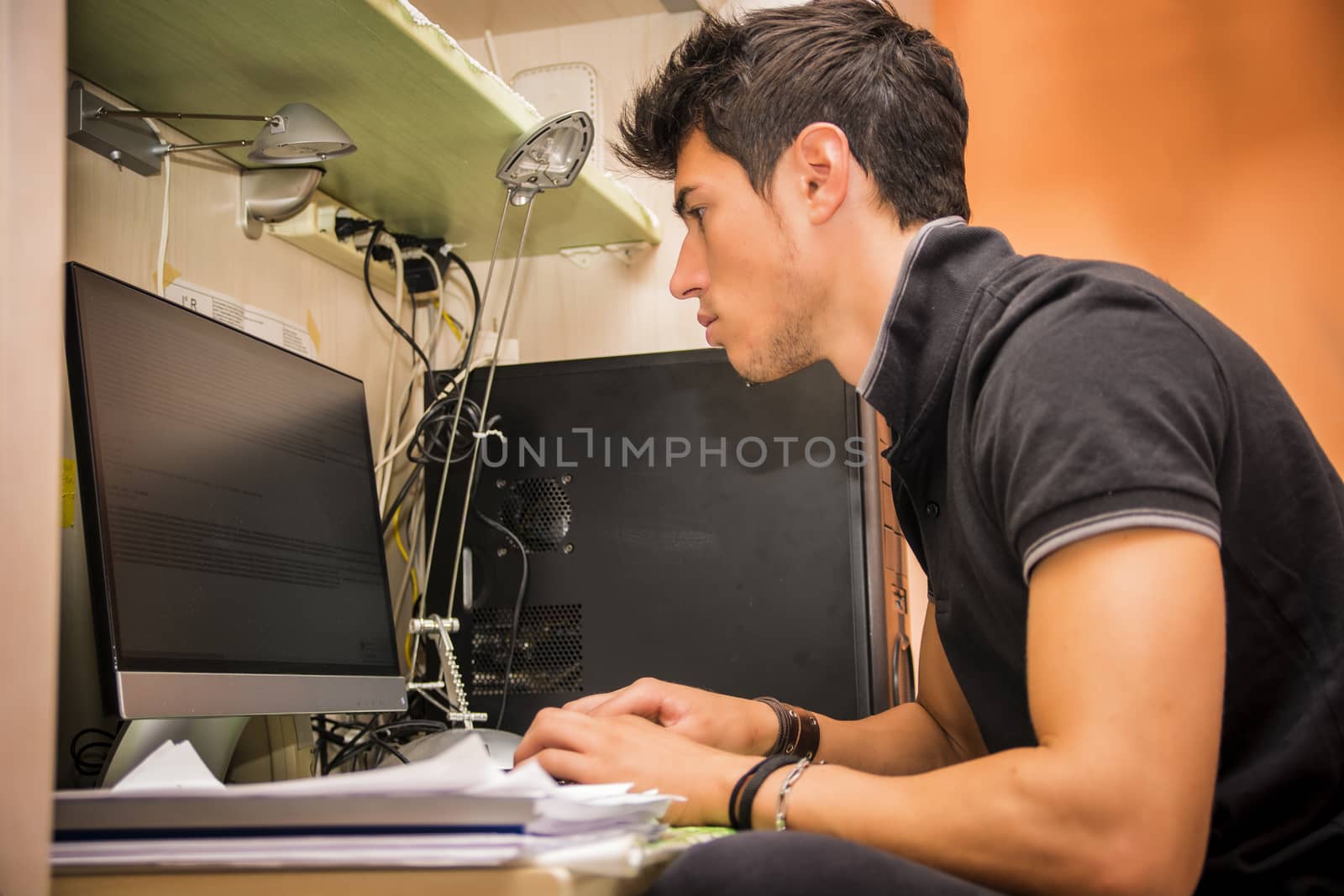 Waist Up Profile of Young Attractive Man with Dark Hair, Sitting at Computer Desk Working on Paper Homework or on His Start-up Business, in Dorm Room