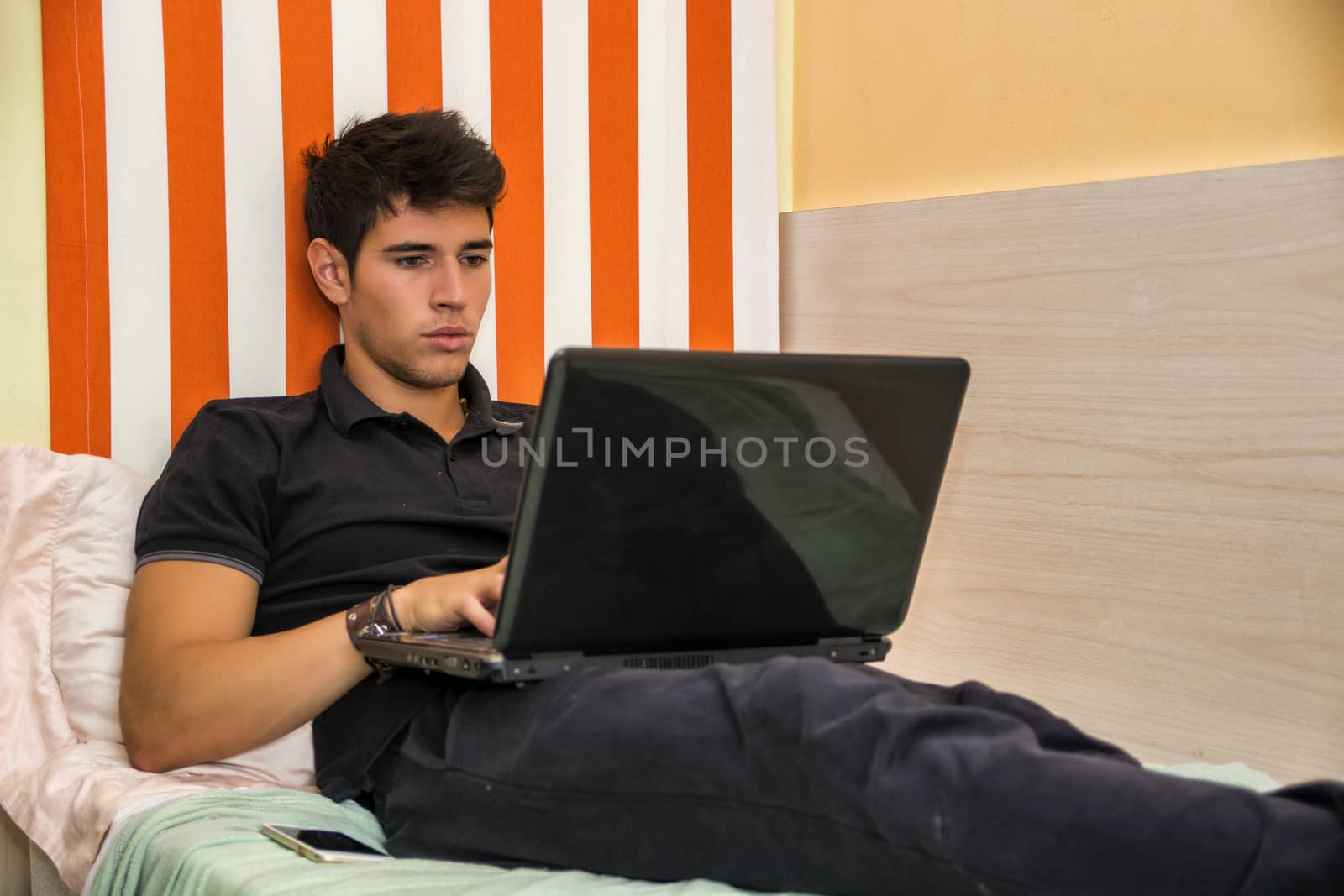Attractive Young Man with Serious Expression, Reclining Comfortably with Laptop on Bed Working on his Start-up Business - Young Male College or University Student Doing Homework, in Bedroom