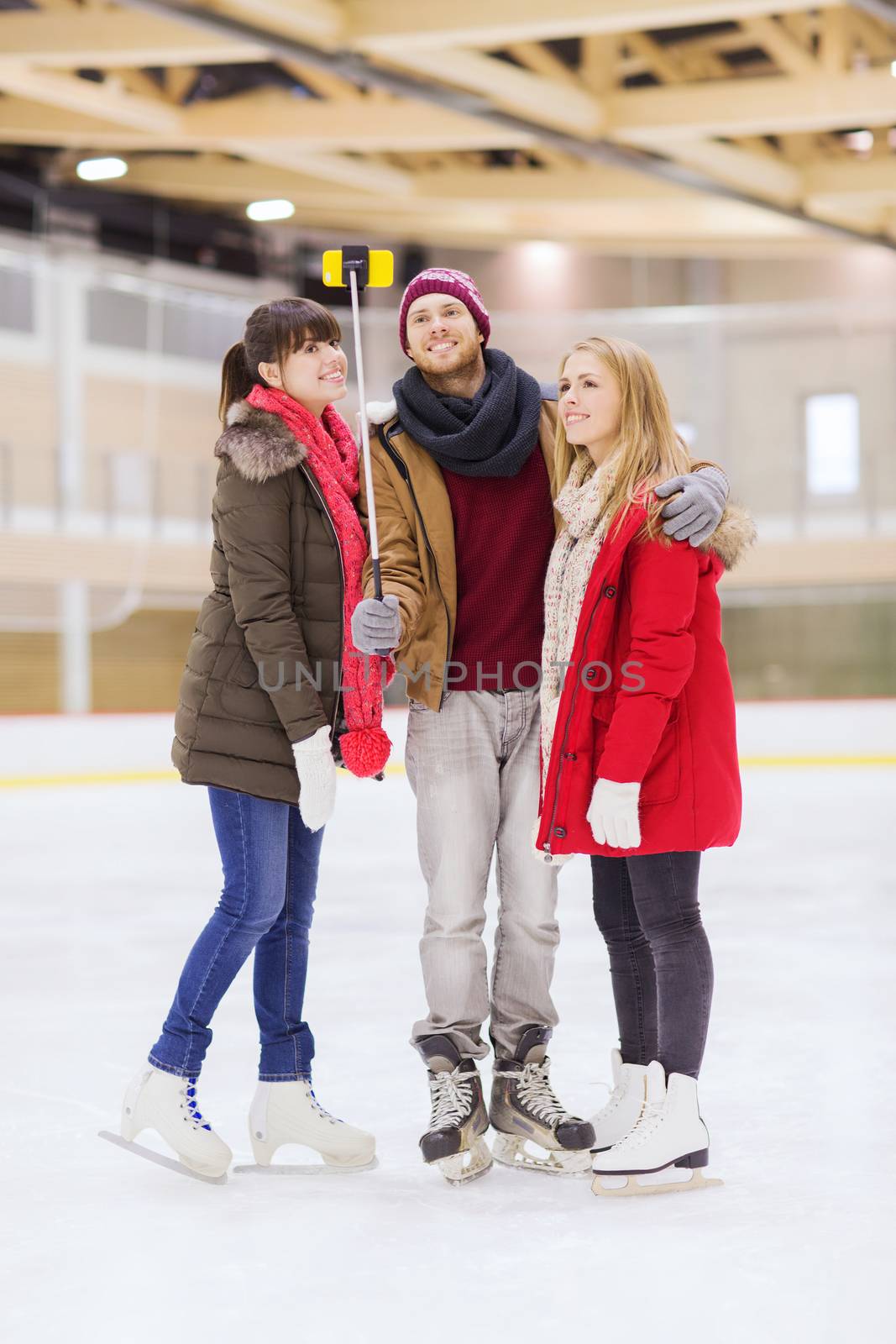 people, friendship, technology and leisure concept - happy friends taking picture with smartphone and selfie stick on skating rink