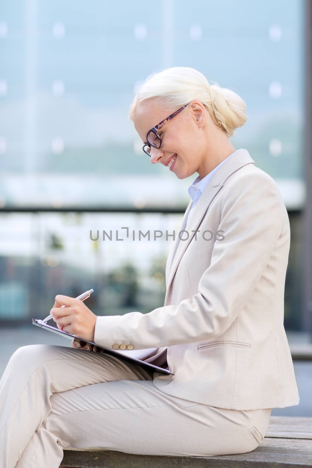 business, people and education concept - young smiling businesswoman in glasses with notepad over office building