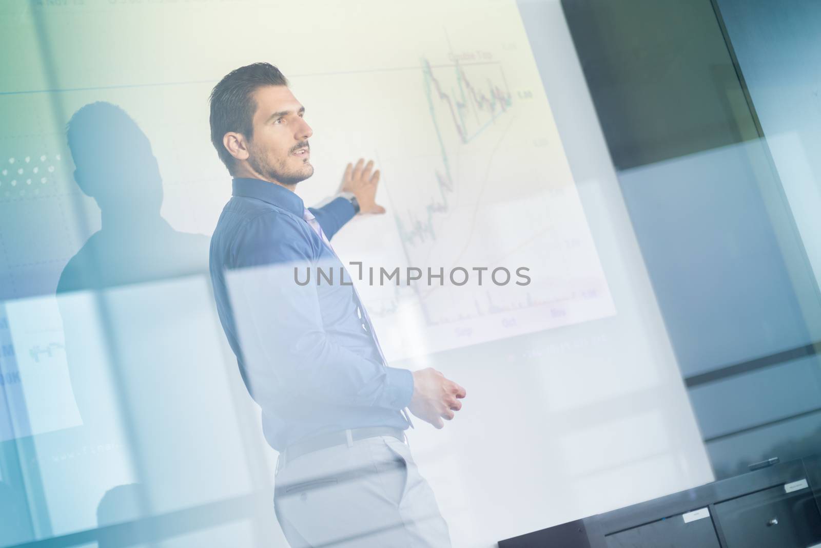 Business man making a presentation in front of whiteboard. Business executive delivering a presentation to his colleagues during meeting or in-house business training. View through glass.