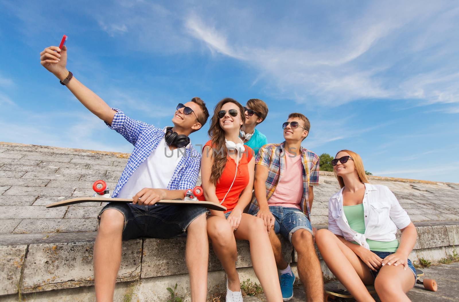 friendship, leisure, summer, technology and people concept - group of smiling friends with skateboard and smartphone making selfie outdoors