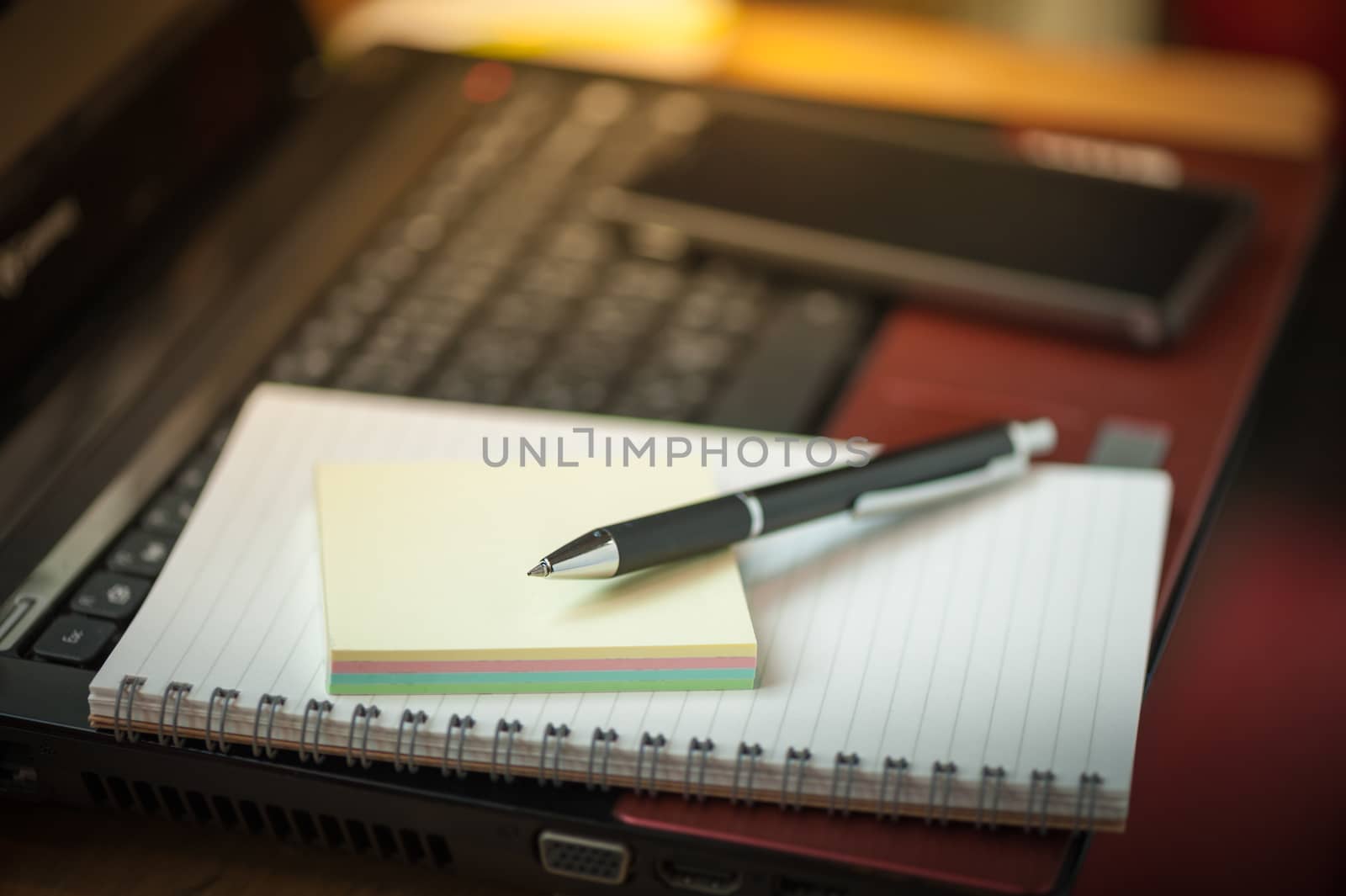 Opened notepad with pen, small sticky paper and black smart phone on laptop computer in coffee shop with morning scene