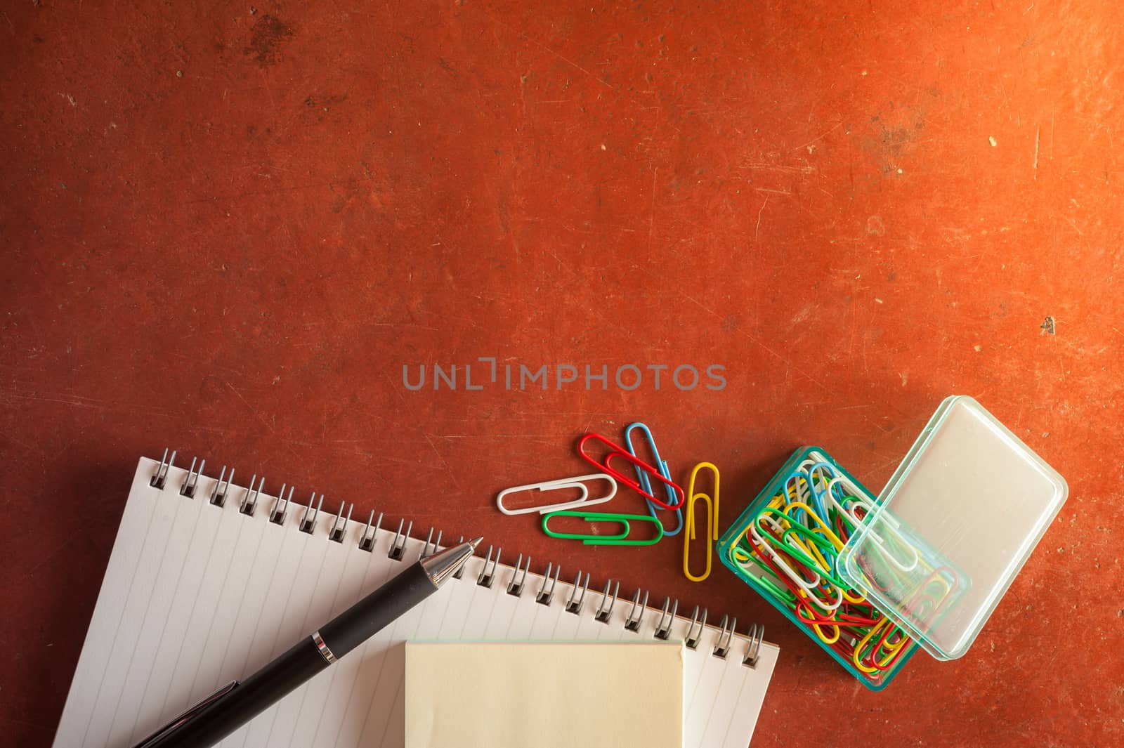 Opened notepad with pen, small sticky paper, and paper clips on red cement background with morning scene