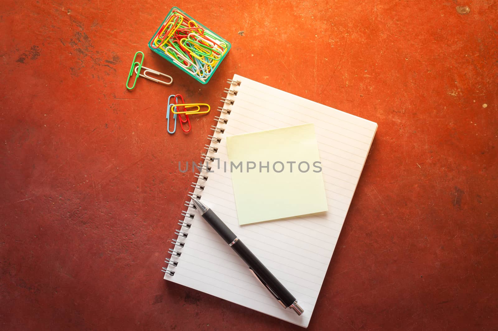 Opened notepad with pen, small sticky paper, and paper clips on red cement background with morning scene