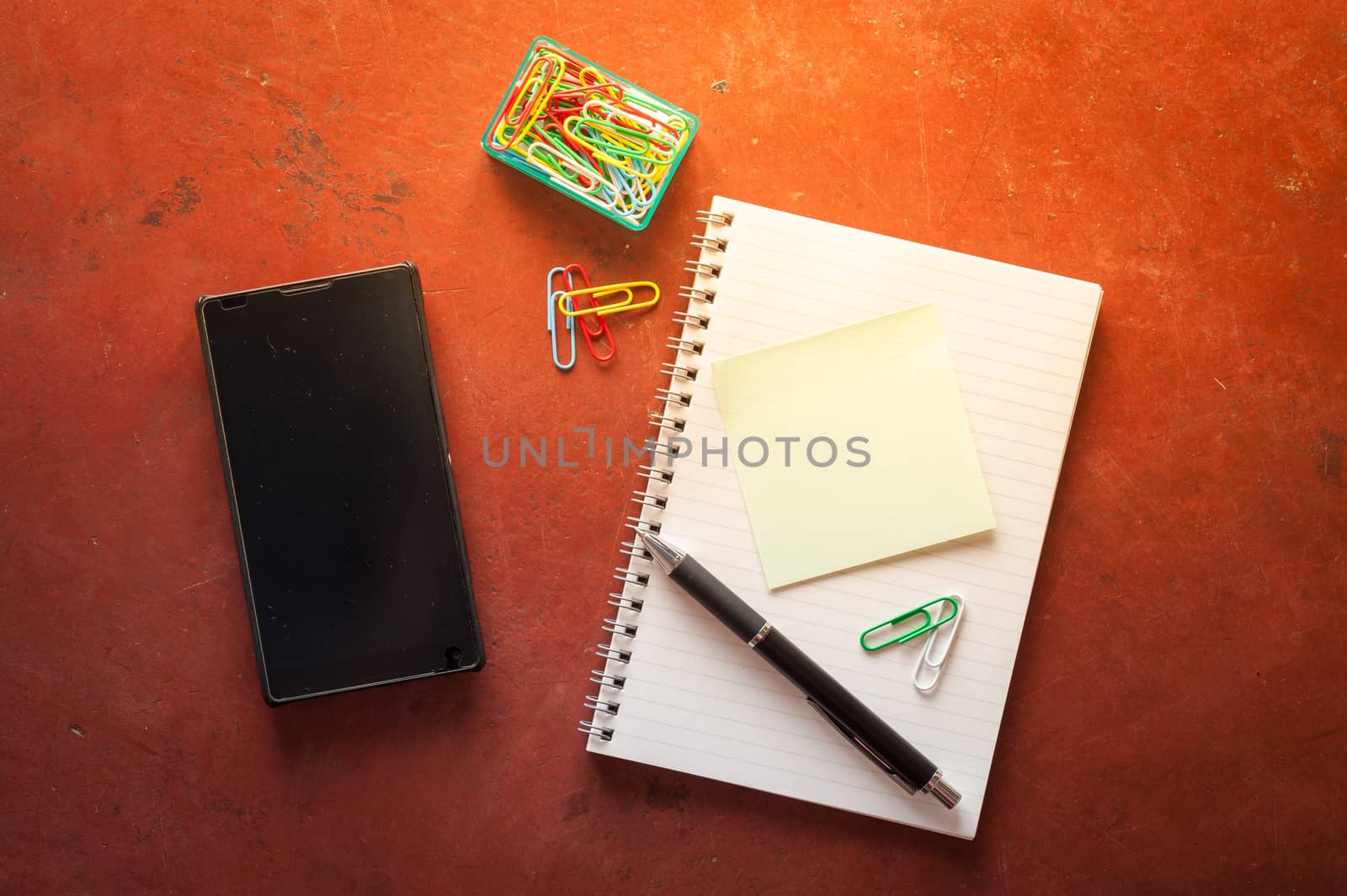 Opened notepad with pen, small sticky paper, smartphone, and paper clips on red cement background with morning scene
