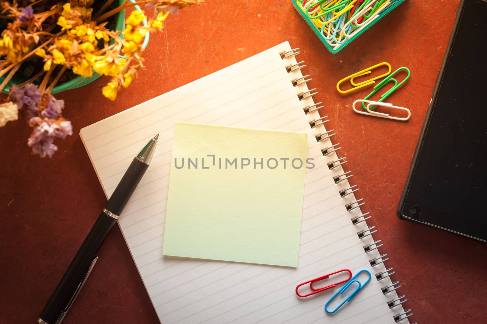 Opened notepad with pen, small sticky paper, smartphone, and paper clips on red cement background with morning scene