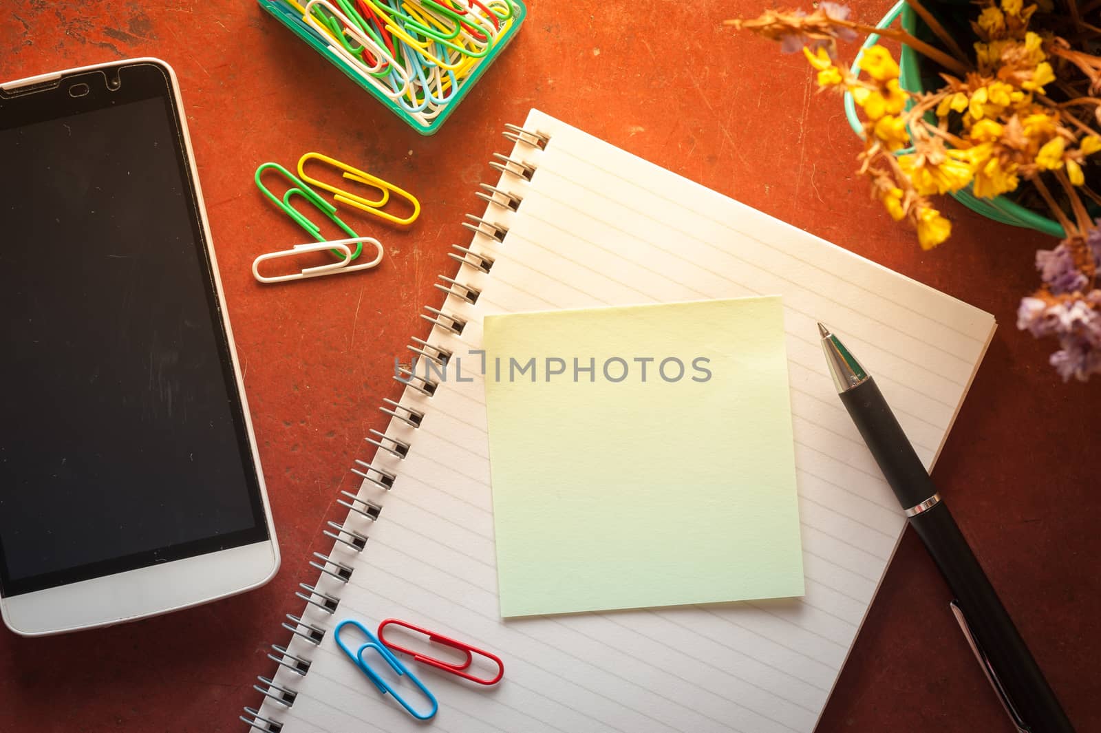 Opened notepad with pen, small sticky paper, smartphone, and paper clips on red cement background with morning scene
