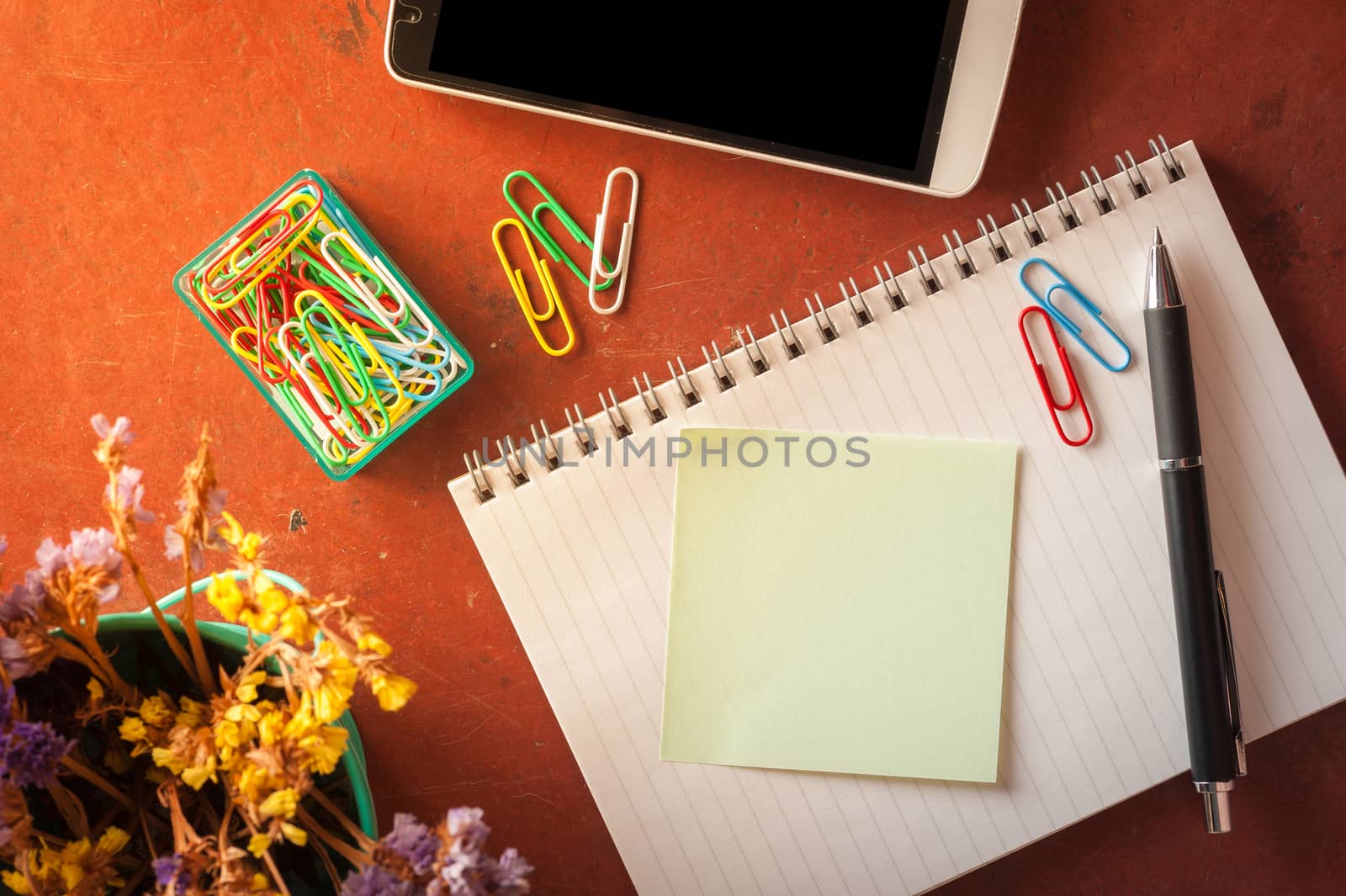 Opened notepad with pen, small sticky paper, smartphone, and paper clips on red cement background with morning scene