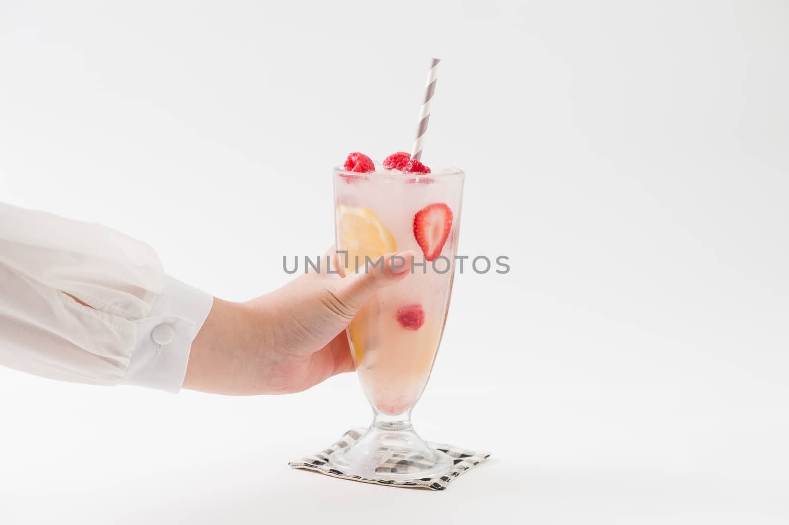 Woman left hand holding glass of iced berry lemonade with striped straw garnished with lemon, raspberry and strawberry