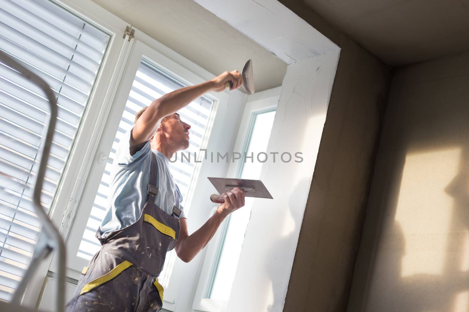 Thirty years old manual worker with wall plastering tools inside a house. Plasterer renovating indoor walls and ceilings with float and plaster.