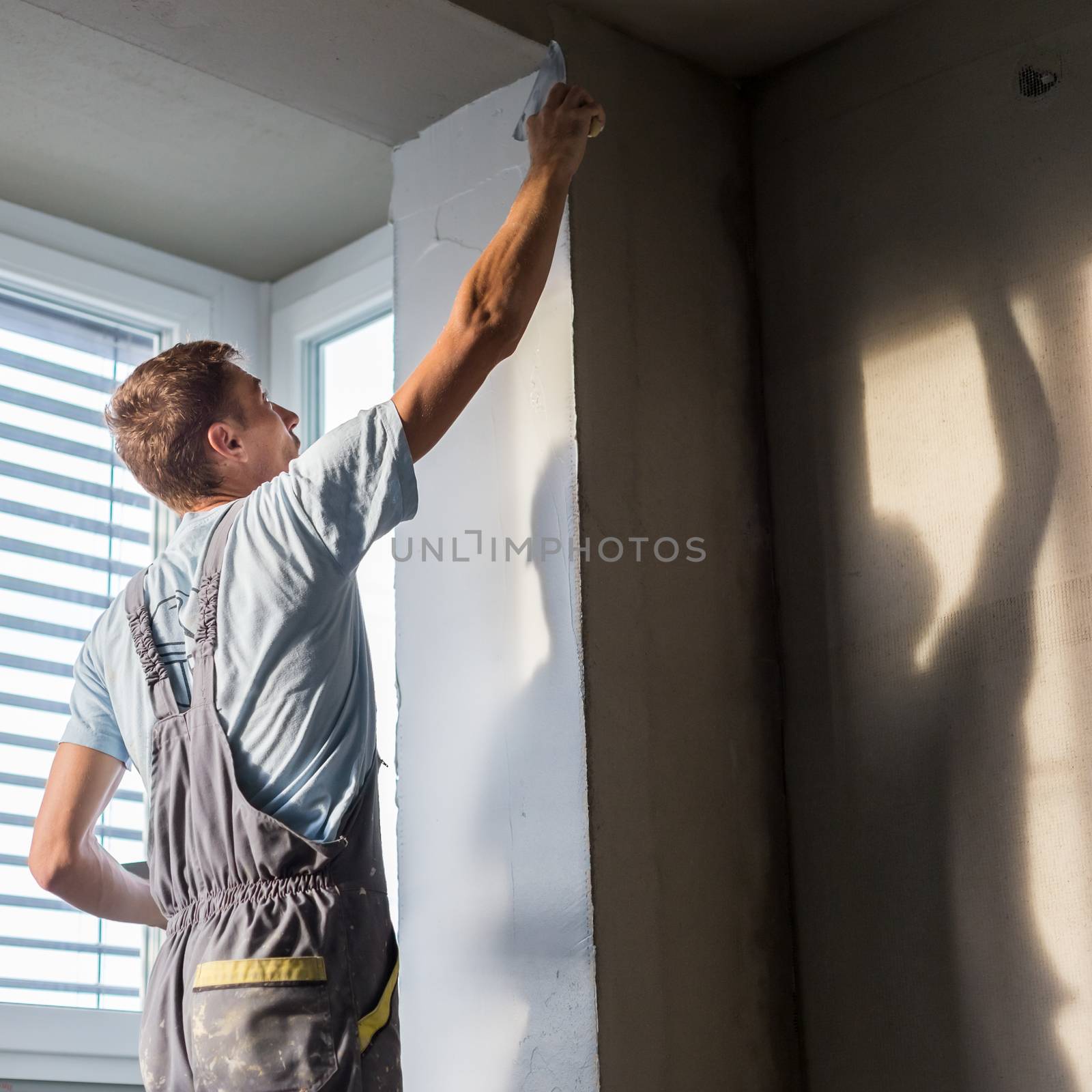 Thirty years old manual worker with wall plastering tools inside a house. Plasterer renovating indoor walls and ceilings with float and plaster.