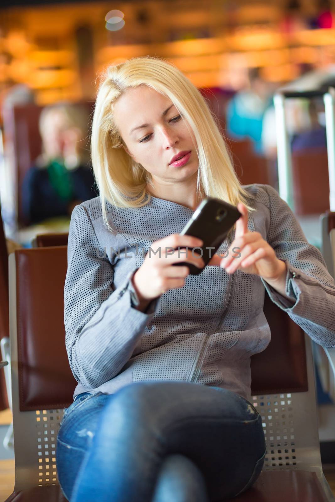 Casual blond young woman using her cell phone while waiting to board a plane at the departure gates. Wireless network hotspot enabling people to access internet conection. Public transport.